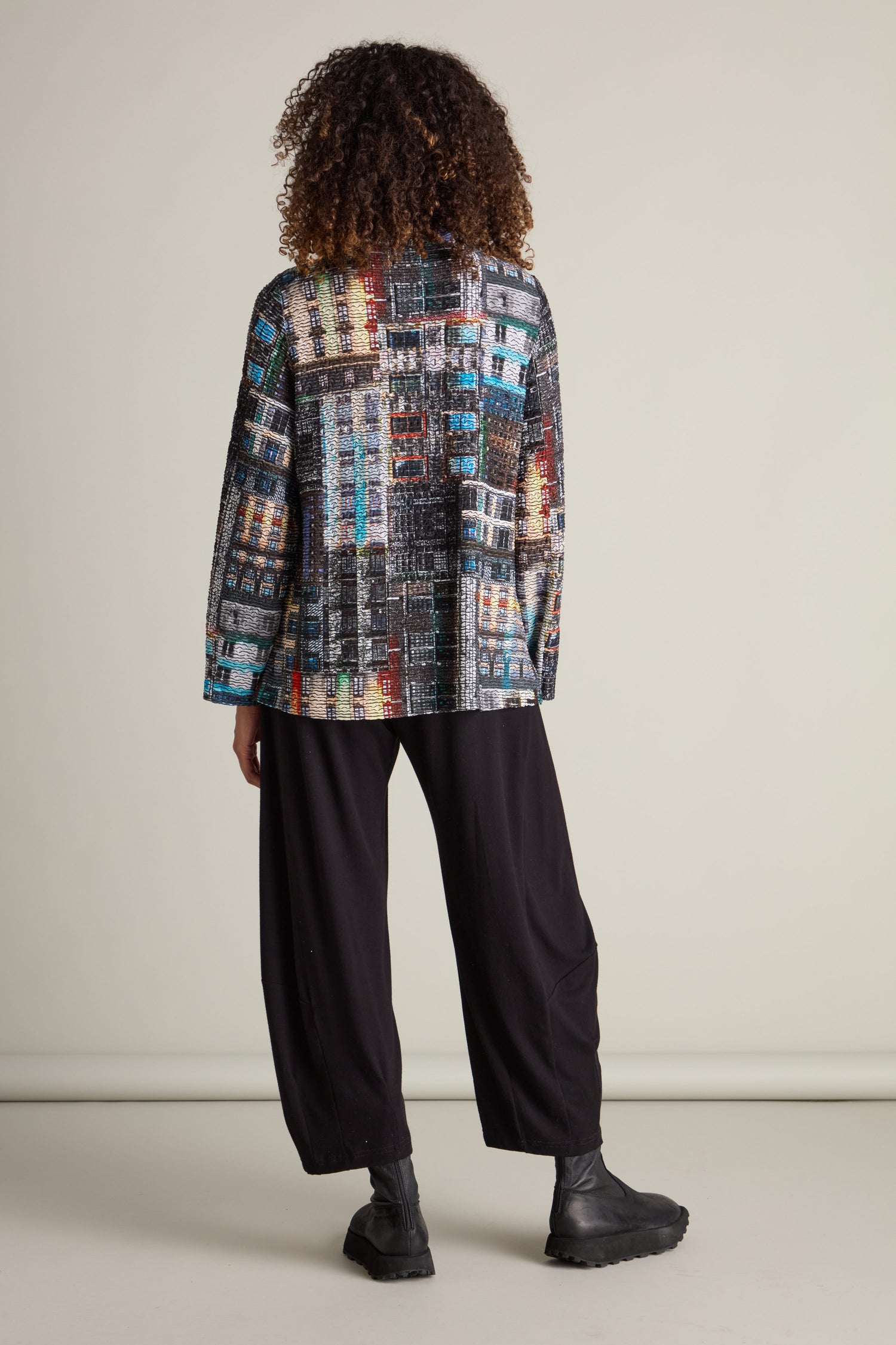 Person with curly hair, wearing a vibrant patterned jacket similar to the Aliza Facade Print Jersey Top, black loose-fitting pants, and black boots, viewed from behind against a plain background.