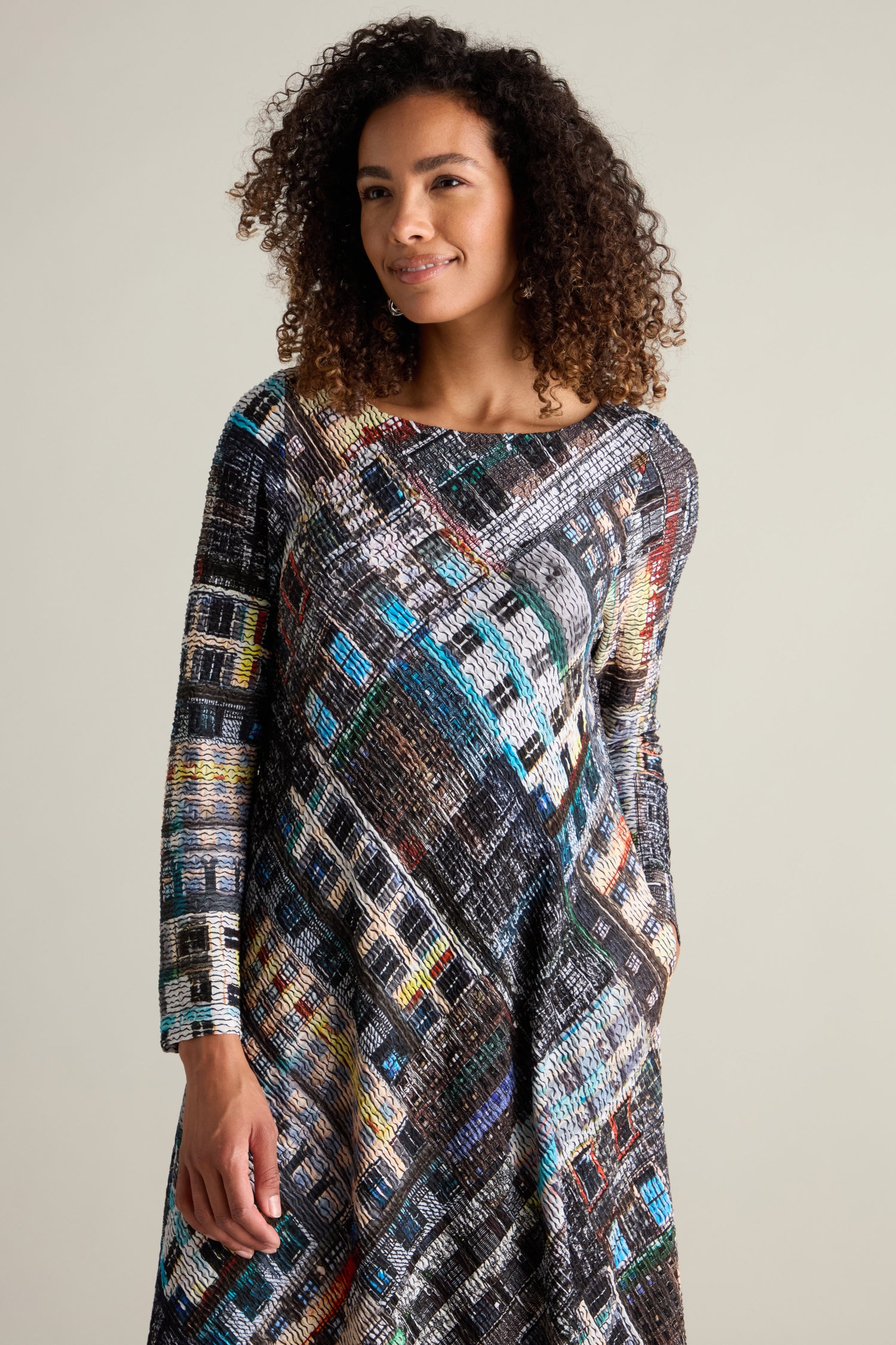 A woman with curly hair wearing the Ramona Facade Print Jersey Dress, featuring long sleeves and a voluminous hem, stands against a light-colored background.