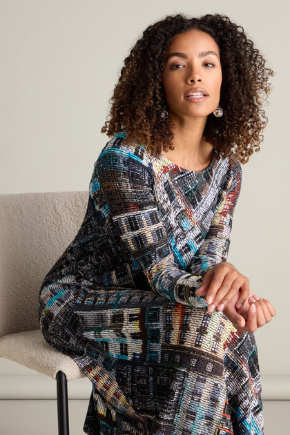 A woman with curly hair is sitting on a chair, wearing the Ramona Facade Print Jersey Dress, which features a house facade print and boasts a voluminous hem.