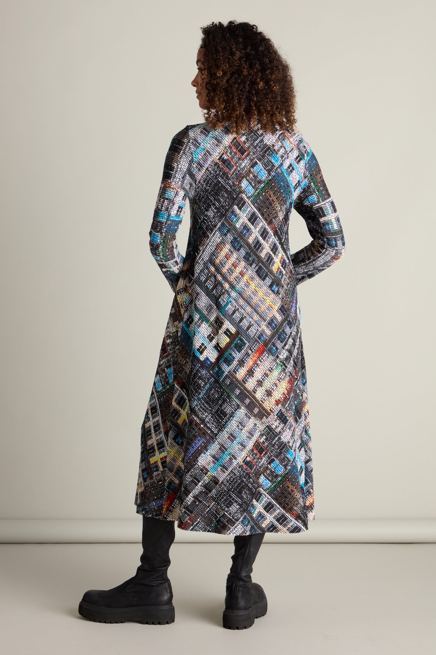 A person with curly hair is standing facing away, wearing the Ramona Facade Print Jersey Dress, which features a long-sleeved boat neckline and colorful geometric House facade design, paired with black boots.