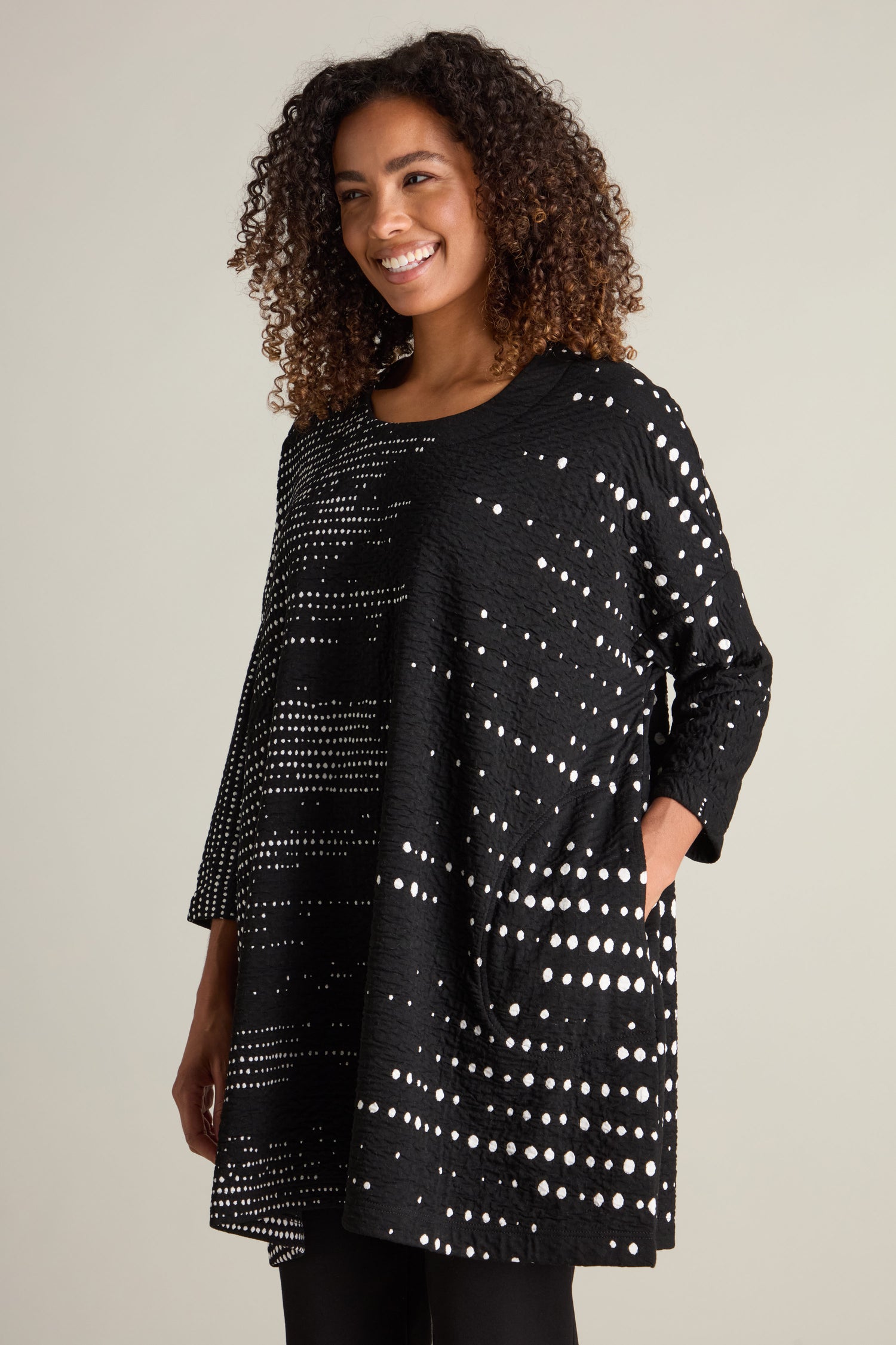 A woman with curly hair is smiling against a plain background, dressed in the Sofia Multi Size Spot Jersey Tunic and with her hands in her pockets. The relaxed silhouette of this piece epitomizes the charm of ecological fashion.