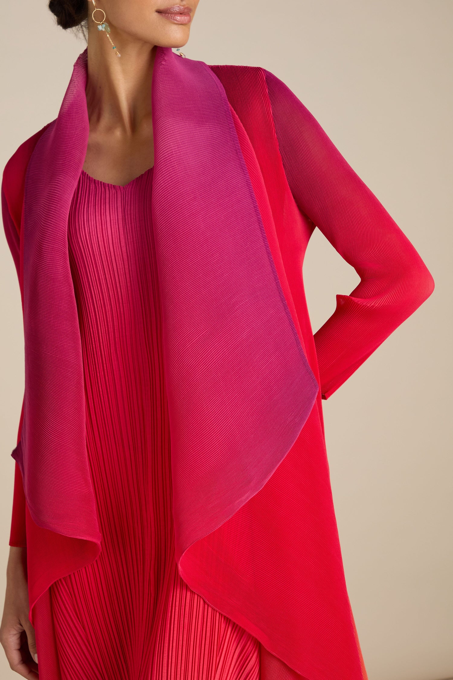 A woman is wearing a red and pink gradient shawl over a red pleated jacket, highlighting her dangling earrings. The ensemble exhibits intricate Japanese craftsmanship, adding a unique flair.