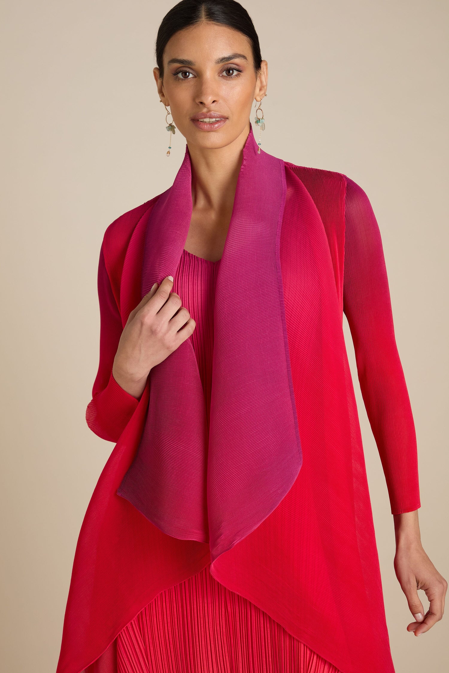 A woman with dark hair wears a Pleated Jacket as part of her long-sleeved, layered red and pink outfit, and is accessorized with dangling earrings. She stands against a plain beige background.