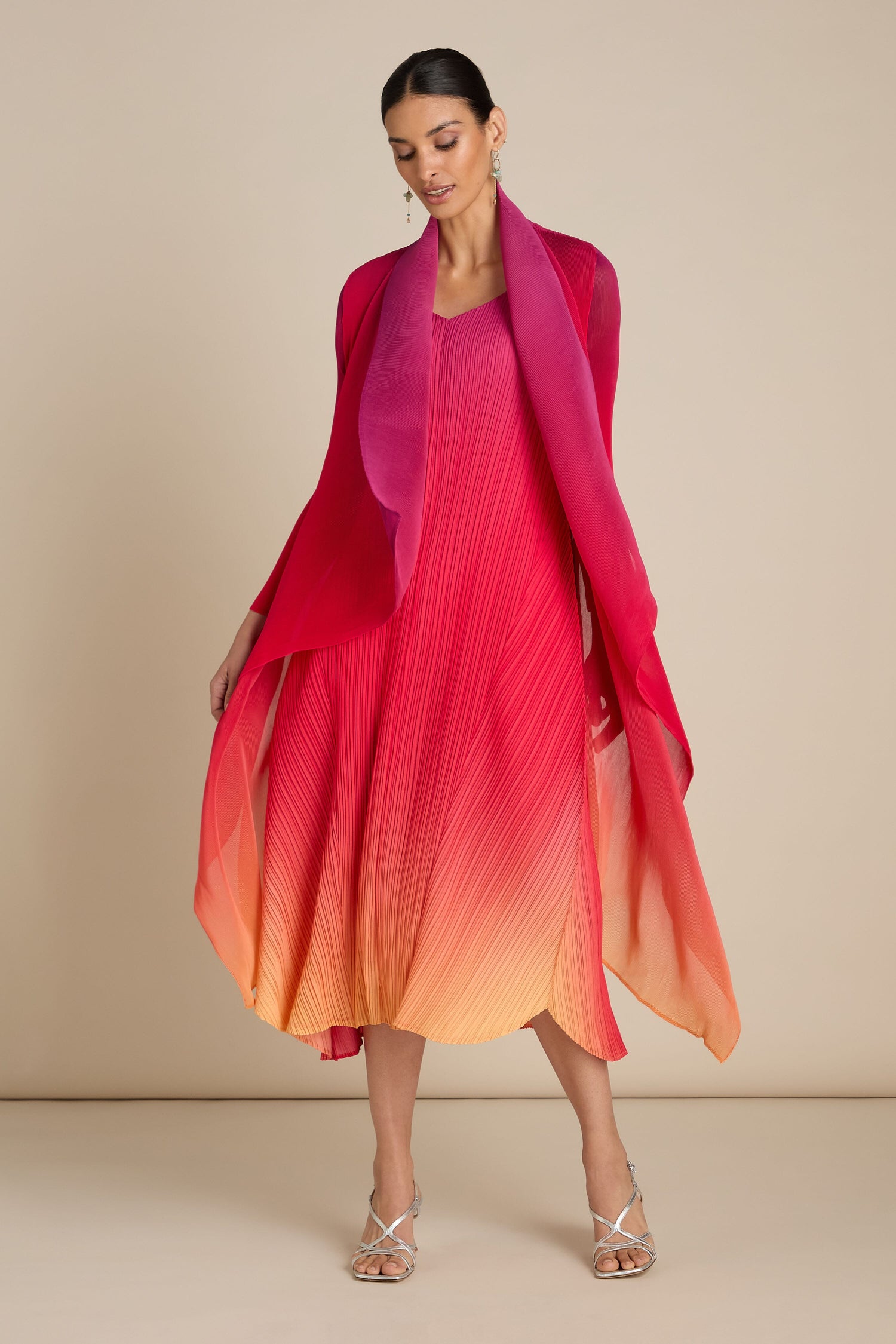 A woman in a gradient red to orange Pleated Jacket with a matching shawl and silver strappy heels stands against a beige background, showcasing the exquisite art of Japanese craft.