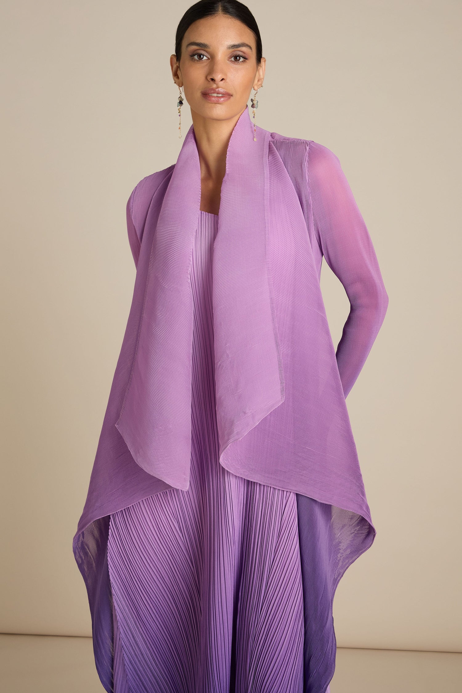 A person stands against a plain background, wearing a flowing, pleated lavender jacket with a matching sheer overlay and dangling earrings that reflect the meticulous attention to detail found in Japanese craft.