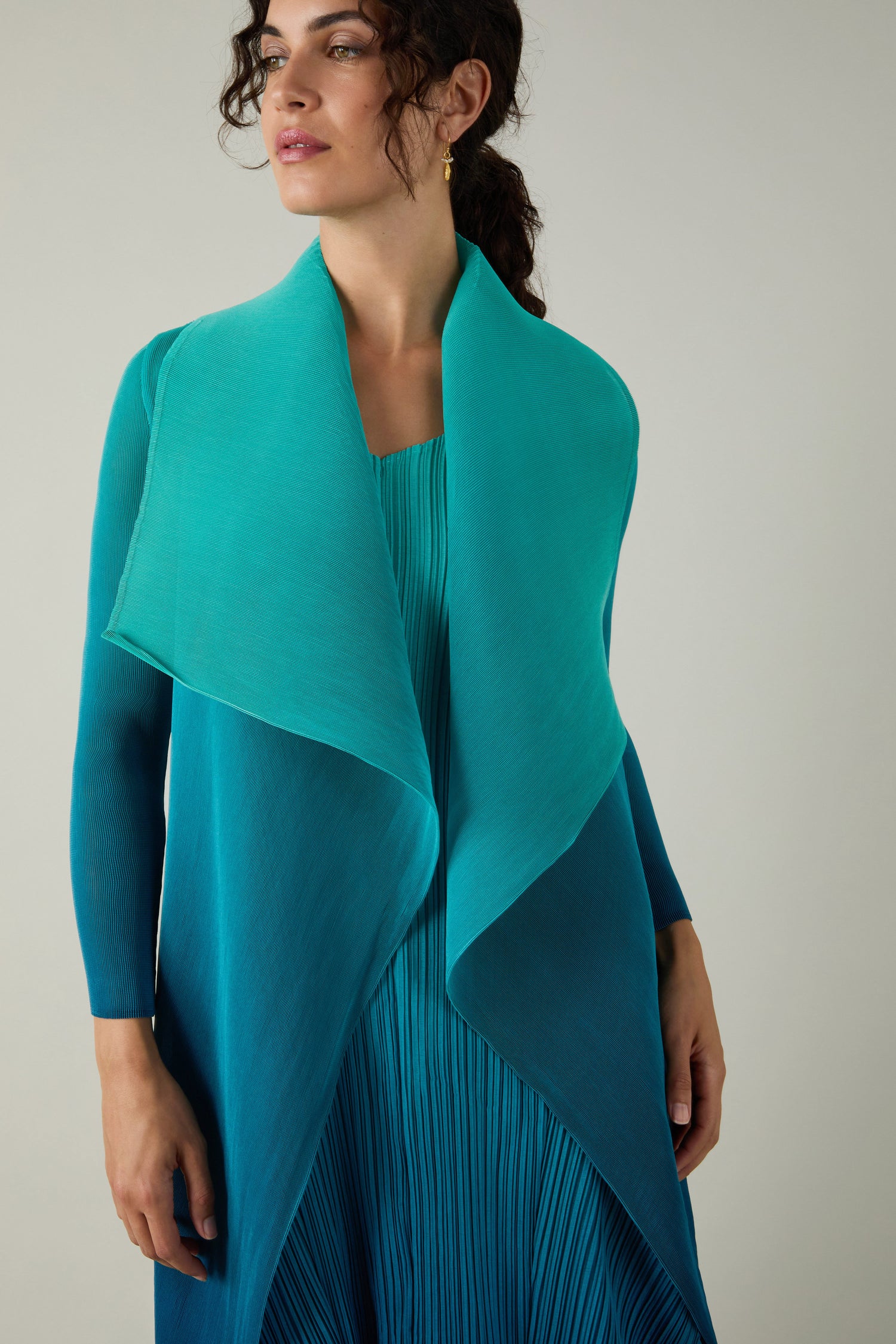 A woman wears a teal Pleated Jacket over a finely pleated dress from an Alquema ensemble, standing against a neutral background.