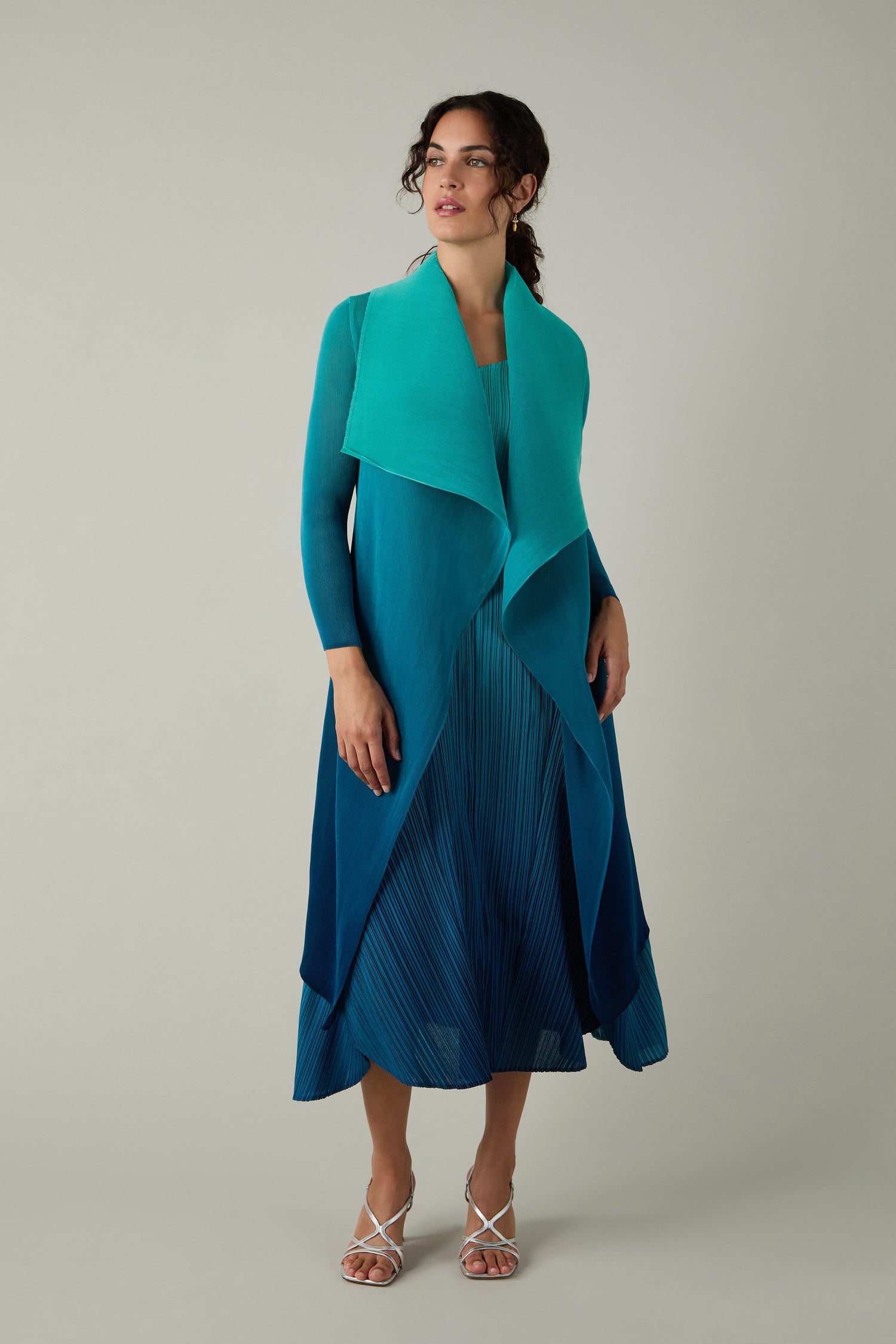 Woman stands wearing the Alquema Pleated Jacket in a teal and blue gradient, paired with a matching teal shawl and white strappy heels, set against a neutral background.