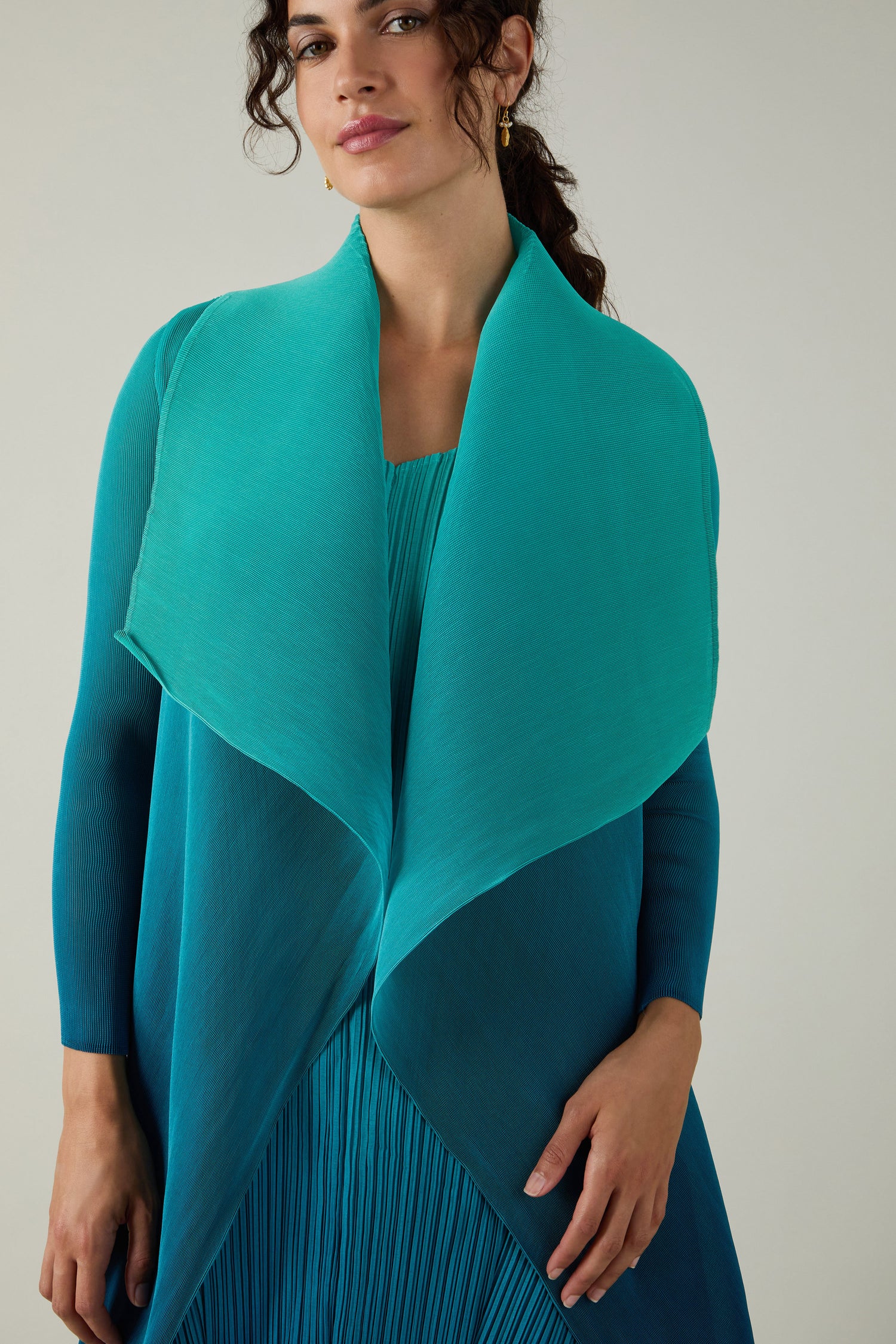 A woman stands against a plain light background, wearing an Alquema Pleated Jacket—a teal draped shawl and blue pleated dress crafted with Japanese shibori techniques.