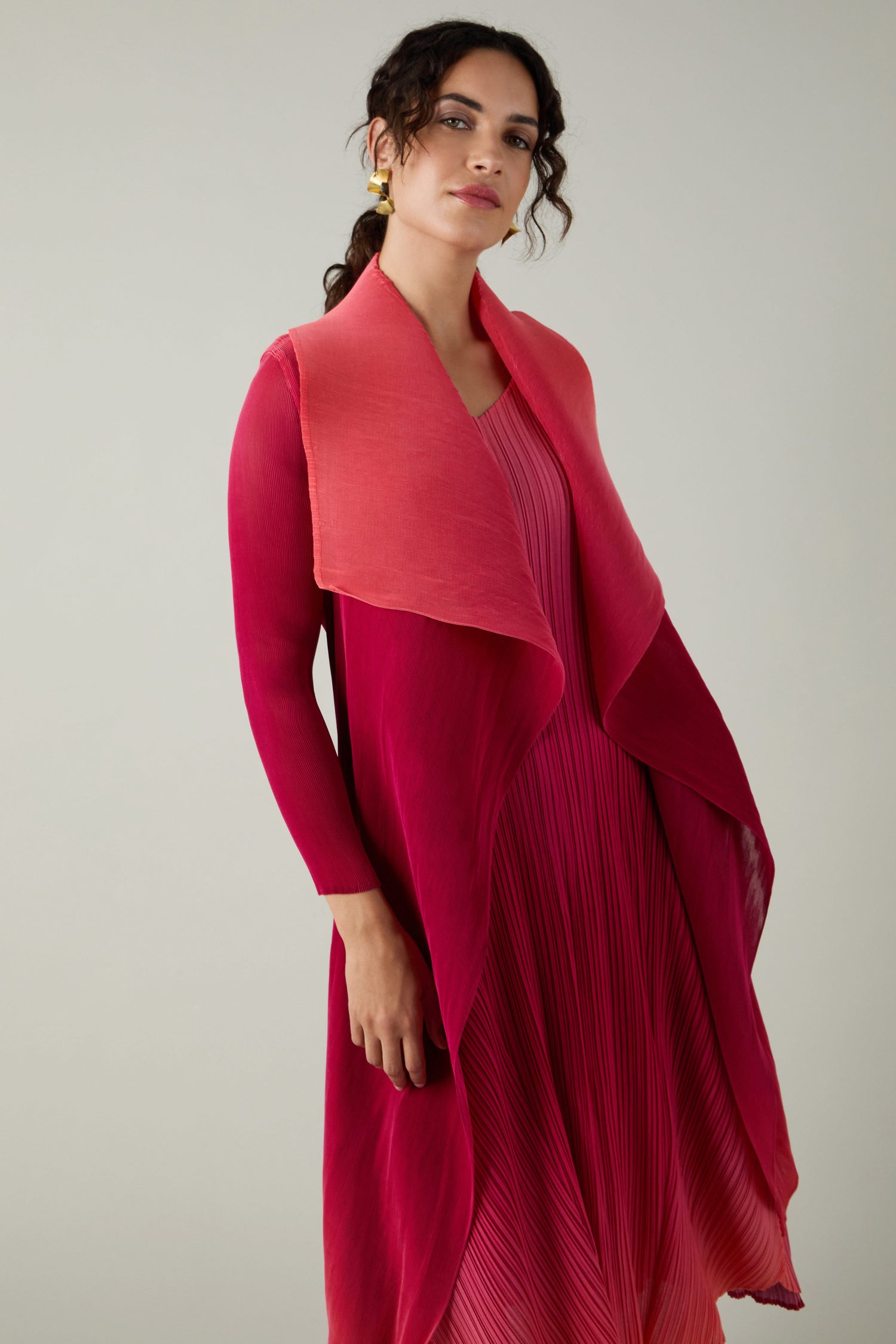 A woman stands against a plain background, wearing Alquema’s Pleated Jacket: a long-sleeved, finely pleated pink and red piece with draped details inspired by Japanese shibori techniques.