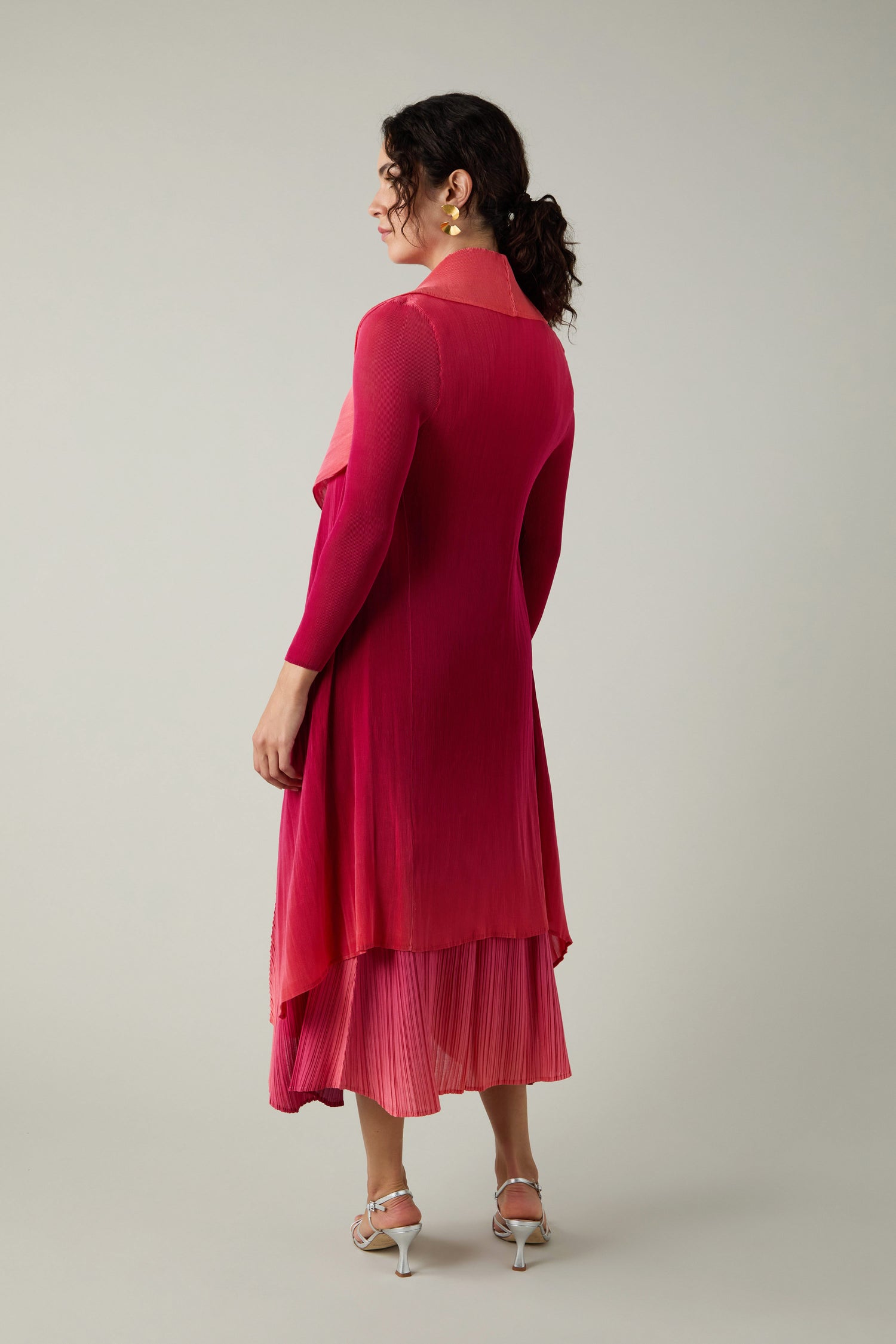 A woman stands with her back to the camera in a finely pleated Alquema Pleated Jacket, layered in pink and red hues, paired with silver heeled sandals against a plain background.