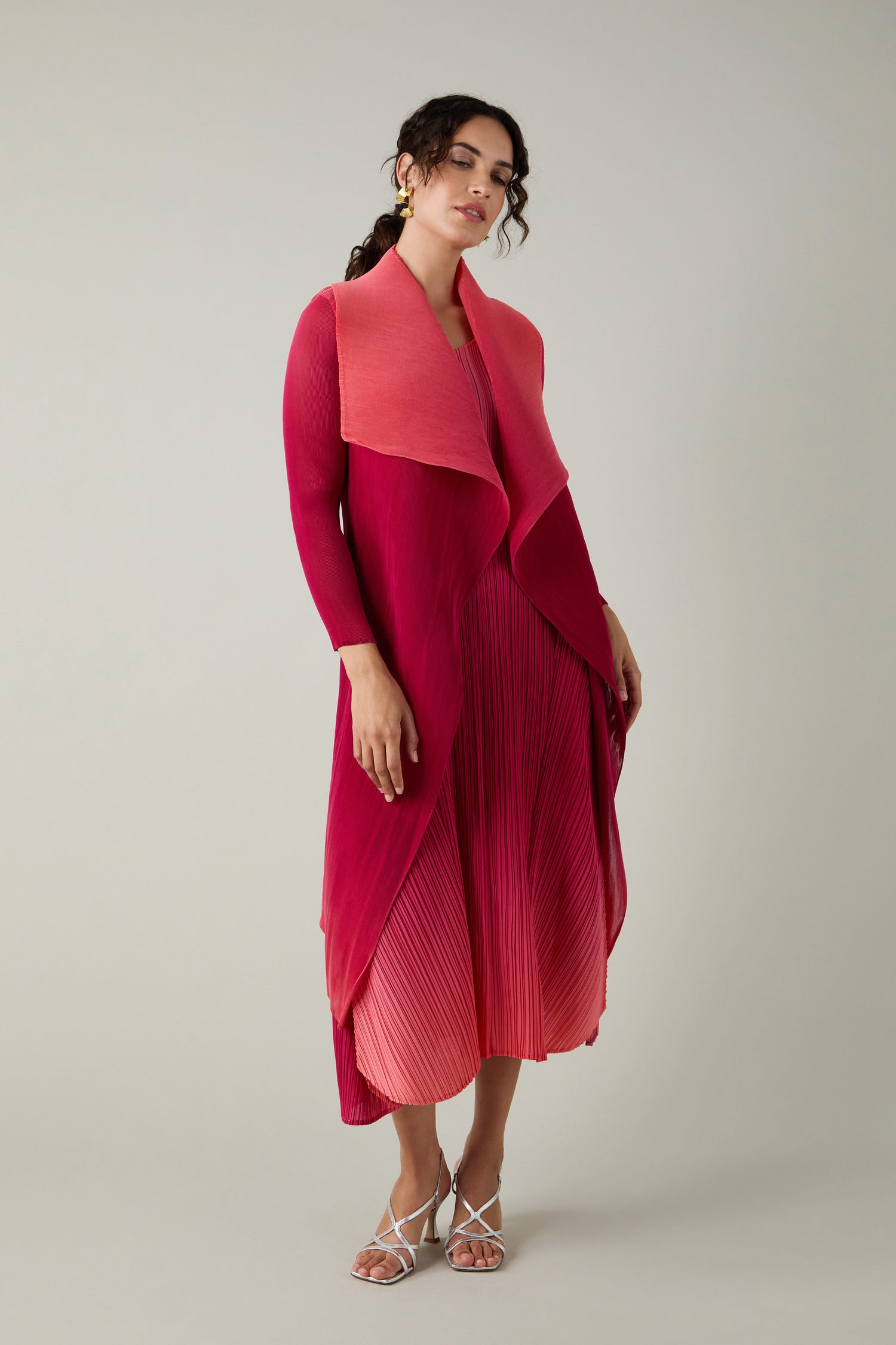 A woman stands in a pink and red ombre Alquema Pleated Jacket ensemble, featuring a finely pleated dress and matching draped cardigan, looking slightly to the side against a plain background.