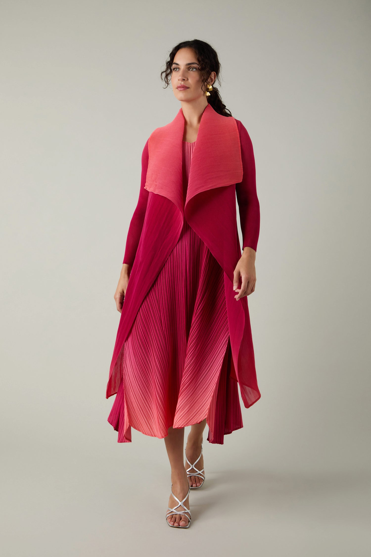 A woman wearing a finely pleated, ombre pink-to-red Pleated Jacket by Alquema with a matching draped vest and white strappy sandals stands against a plain background.