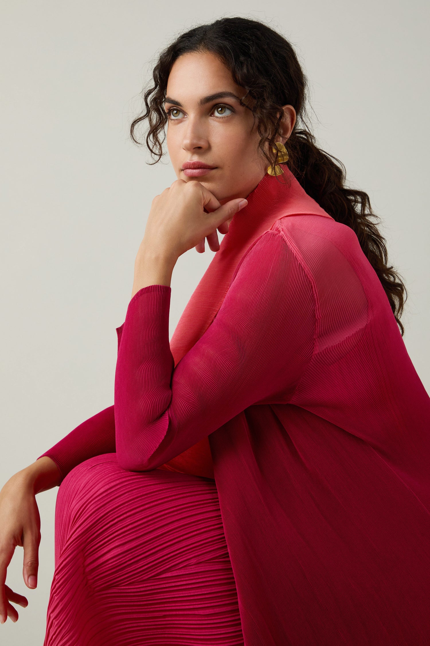 A woman with curly hair, wearing a brightly colored Pleated Jacket inspired by Japanese shibori techniques, sits with her hand on her chin and gazes slightly upward against a plain background.