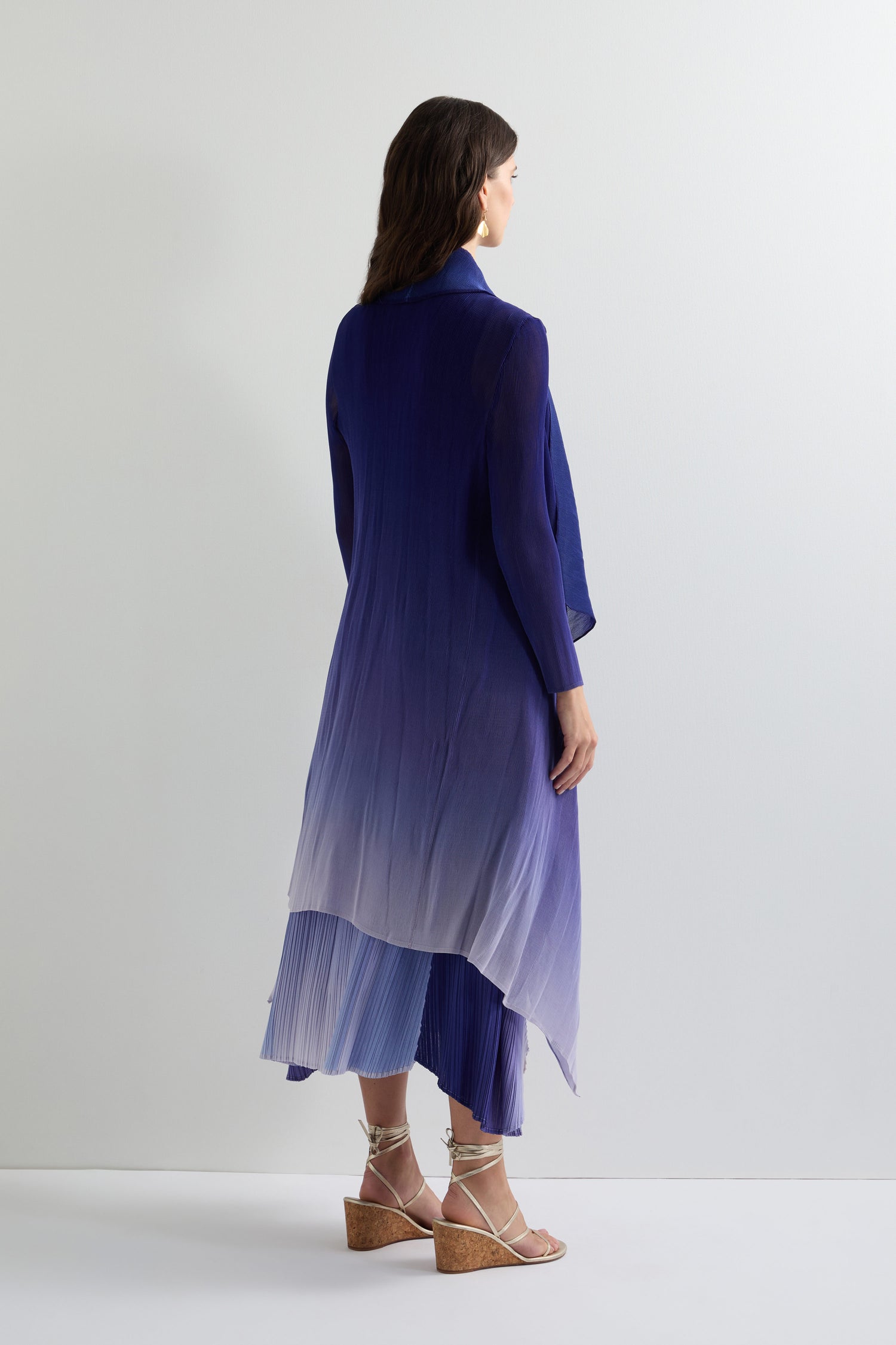 A woman with long brown hair, wearing a Pleated Jacket and sandals, stands against a plain background, facing away. Her look effortlessly blends contemporary elegance with simplicity.