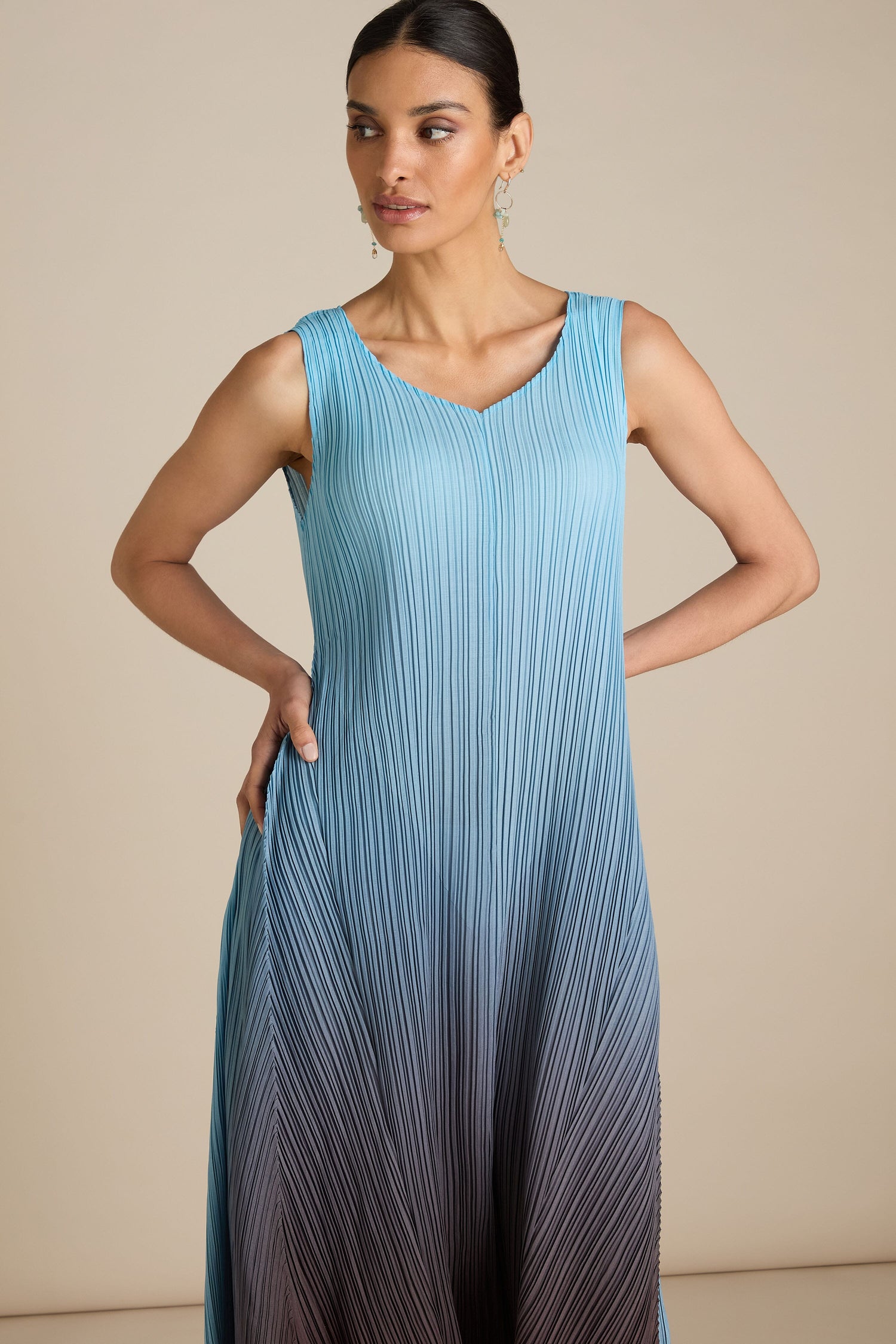 A woman wearing the Pleated Dress, which features a sleeveless blue-to-gray gradient and a relaxed silhouette, poses against a neutral background. With her hair tied back, she gazes slightly to the side with effortless elegance.