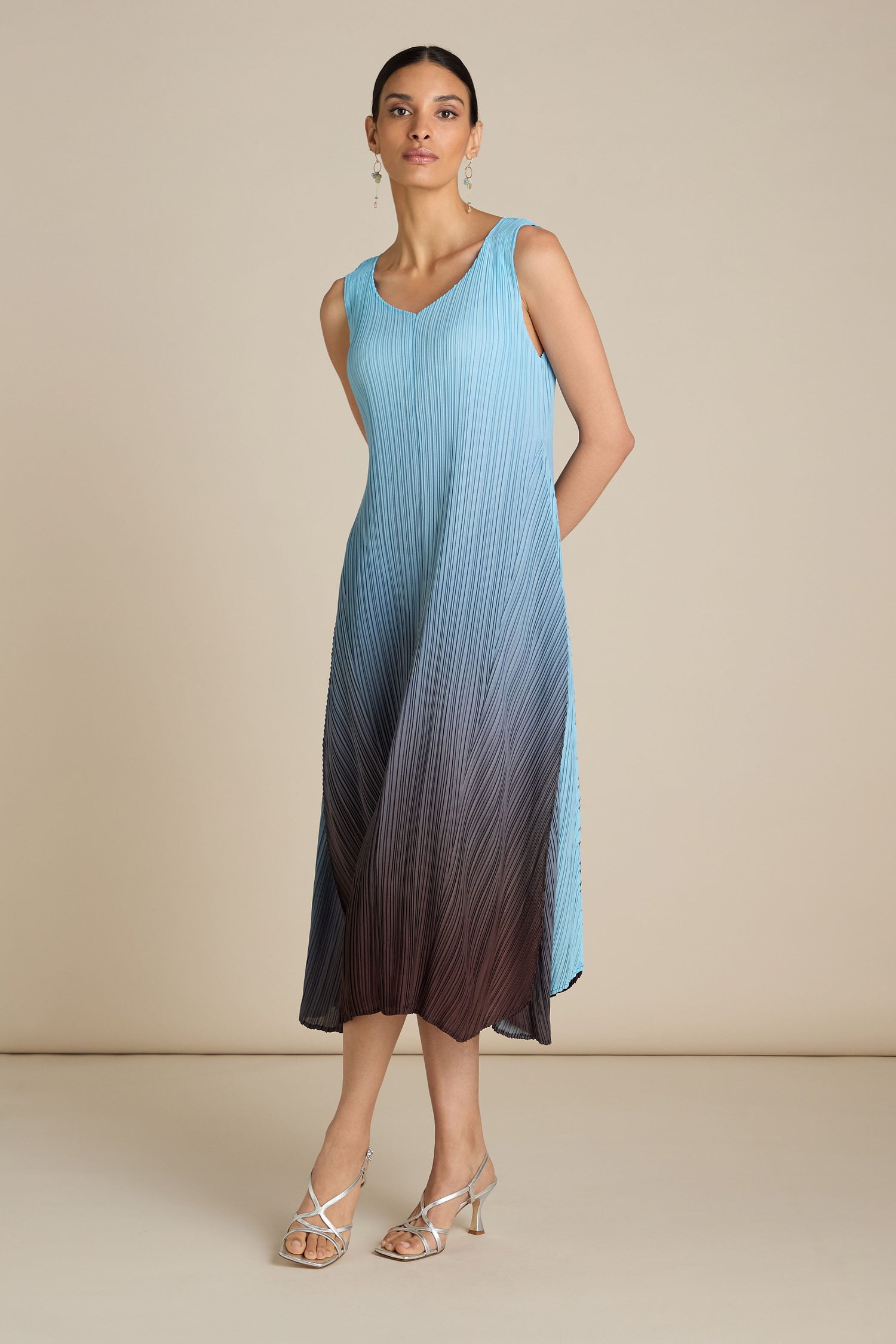 Person wearing a sleeveless, gradient blue-to-brown Pleated Dress stands against a plain background with hands behind back. The relaxed silhouette adds a touch of bohemian flair, accessorized with drop earrings and strappy heels.