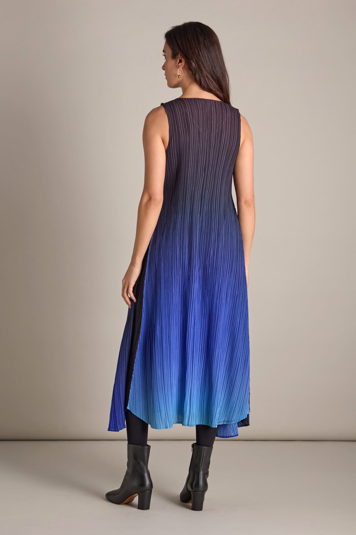 A woman stands with her back turned in a pleated dress, showcasing its exquisite fabric that transitions from dark to light blue, complemented by black ankle boots. The outfit gently captures a bohemian essence.