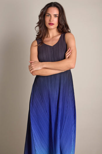 A person stands with arms crossed, dressed in a sleeveless Pleated Dress showcasing a gradient of blue hues and intricate pleats, offering a bohemian touch.