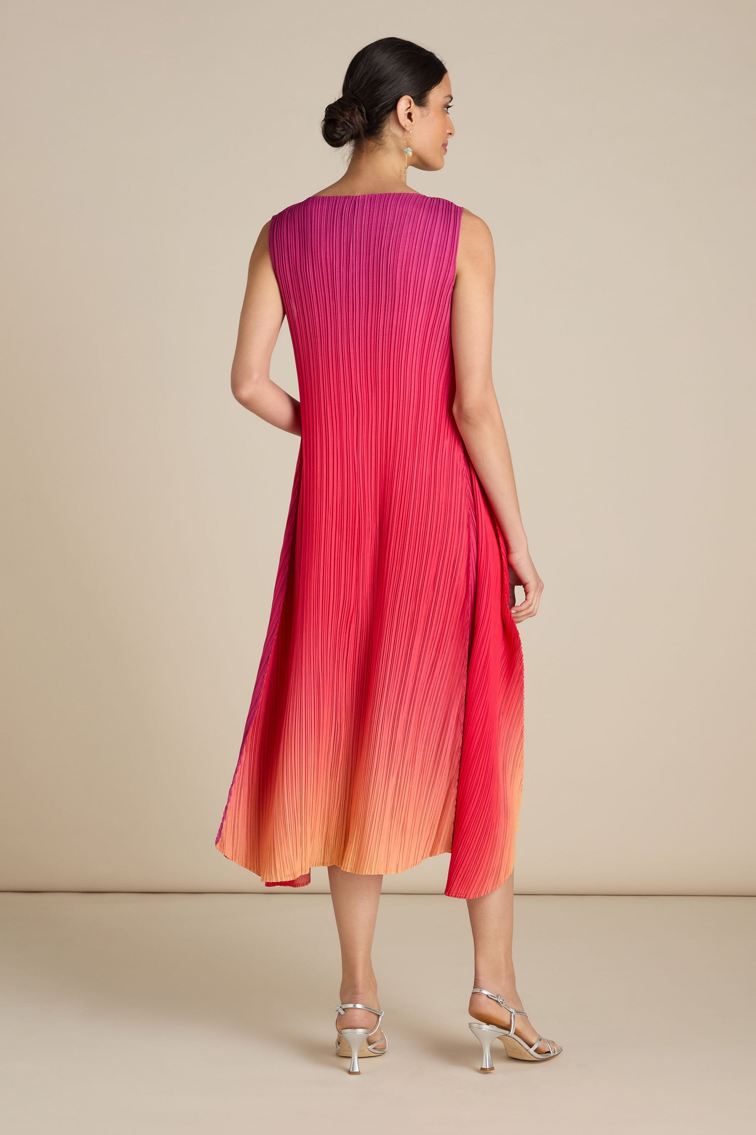 A woman in the Pleated Dress, featuring a sleeveless design and a gradient from pink to orange, stands facing away, highlighting its relaxed silhouette. Her hair is styled in a low bun, and she complements her outfit with silver heeled sandals, giving off a bohemian vibe.