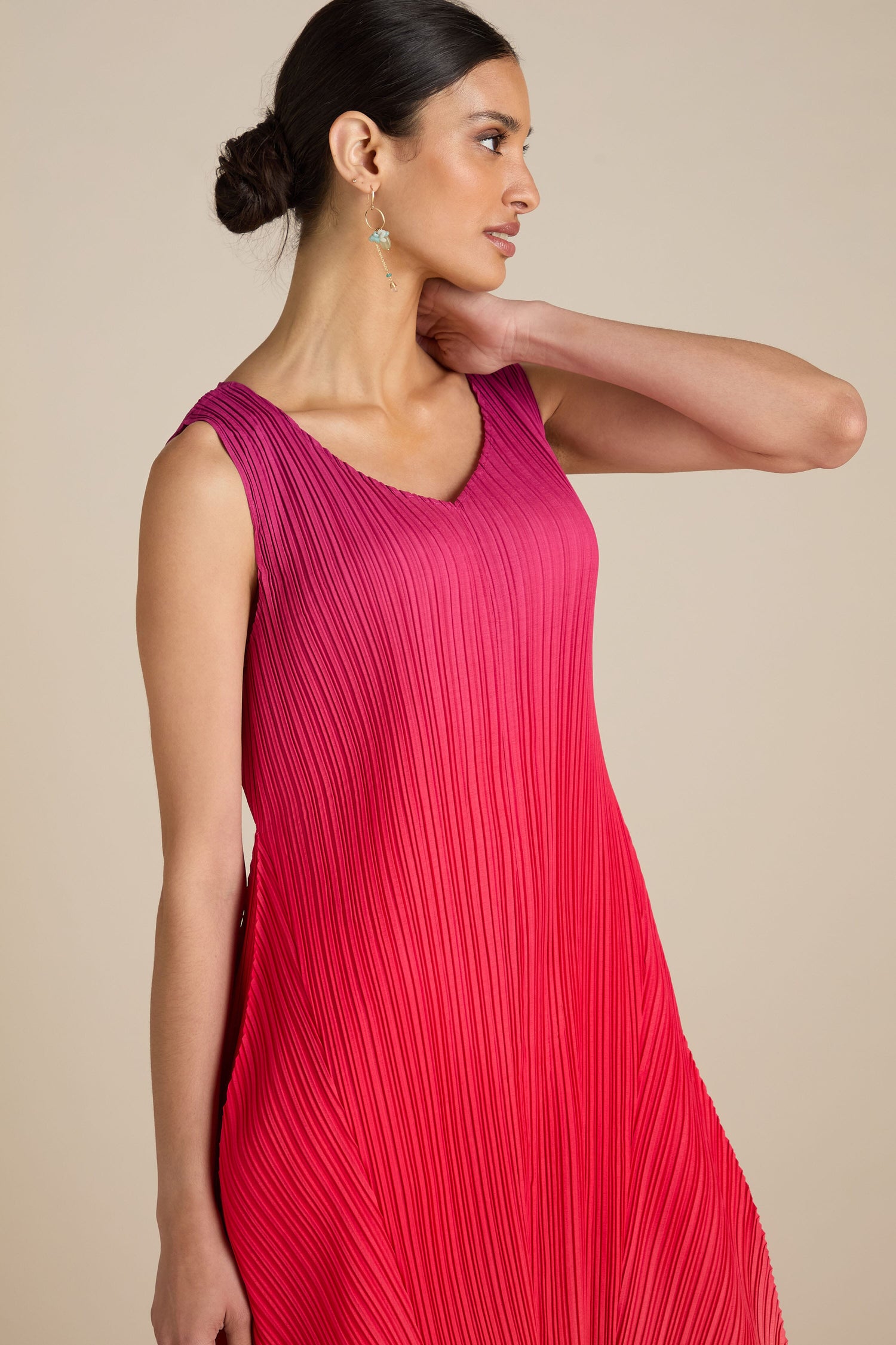 A woman in a sleeveless Pleated Dress featuring a red gradient and a relaxed silhouette, her hair tied back, looks to the side against a beige background.