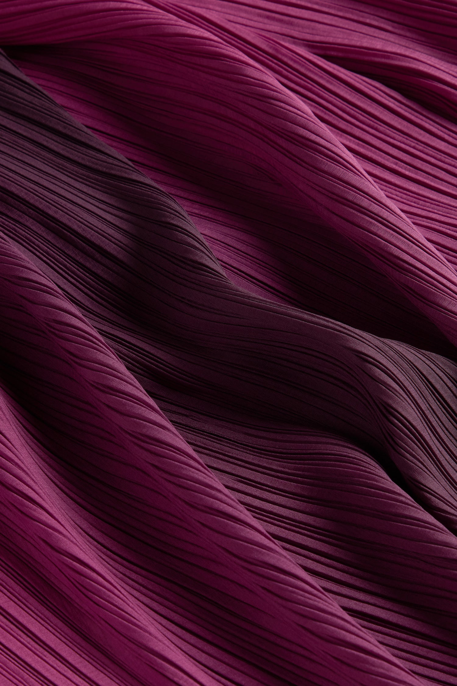 A close-up of the Pleated Dress features fabric in shades of magenta and dark purple, highlighting its intricate texture and wave-like patterns that evoke a bohemian flair.