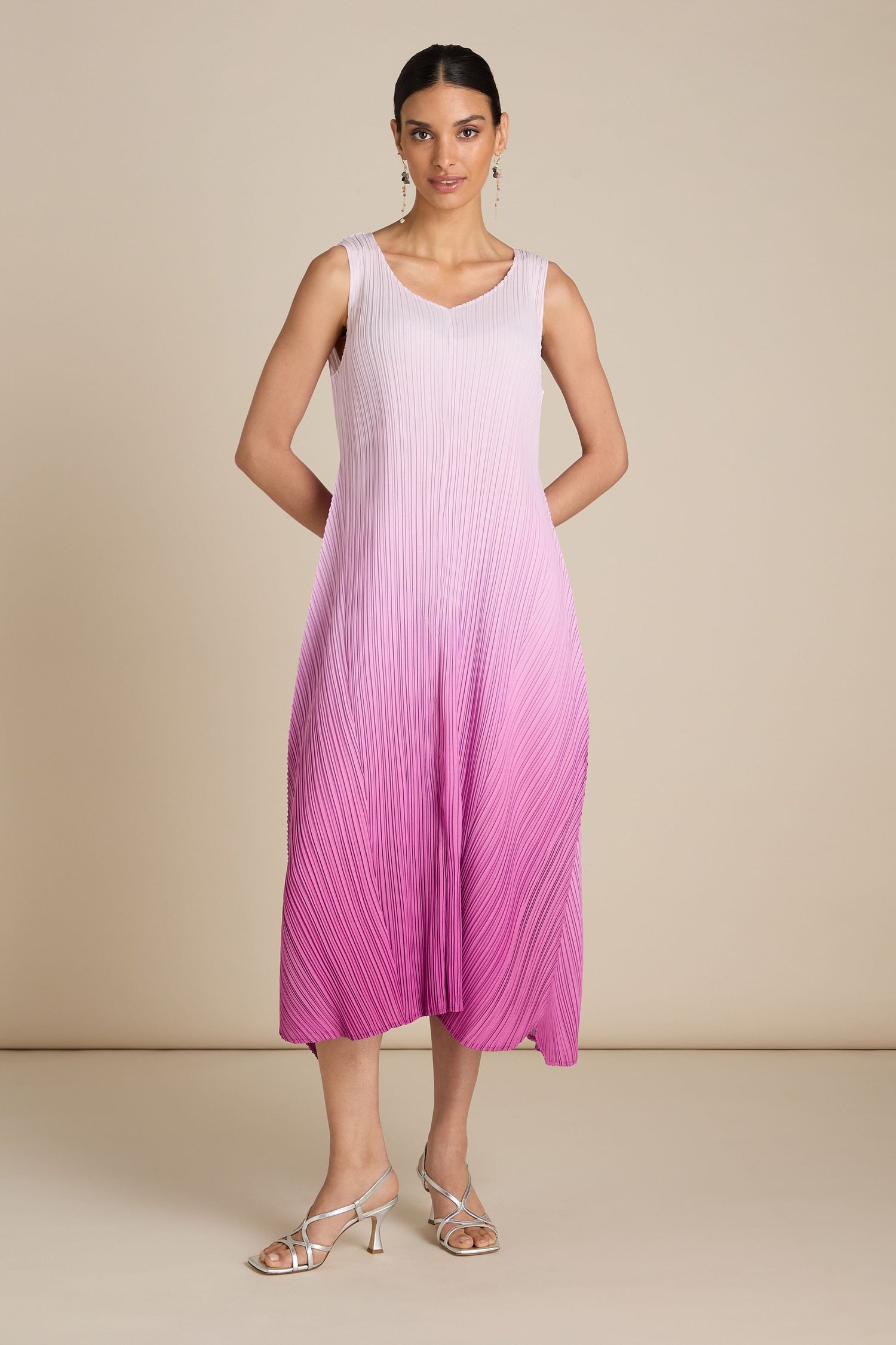 A woman stands in a studio wearing a sleeveless, gradient pink Pleated Dress with a relaxed silhouette and silver sandals.