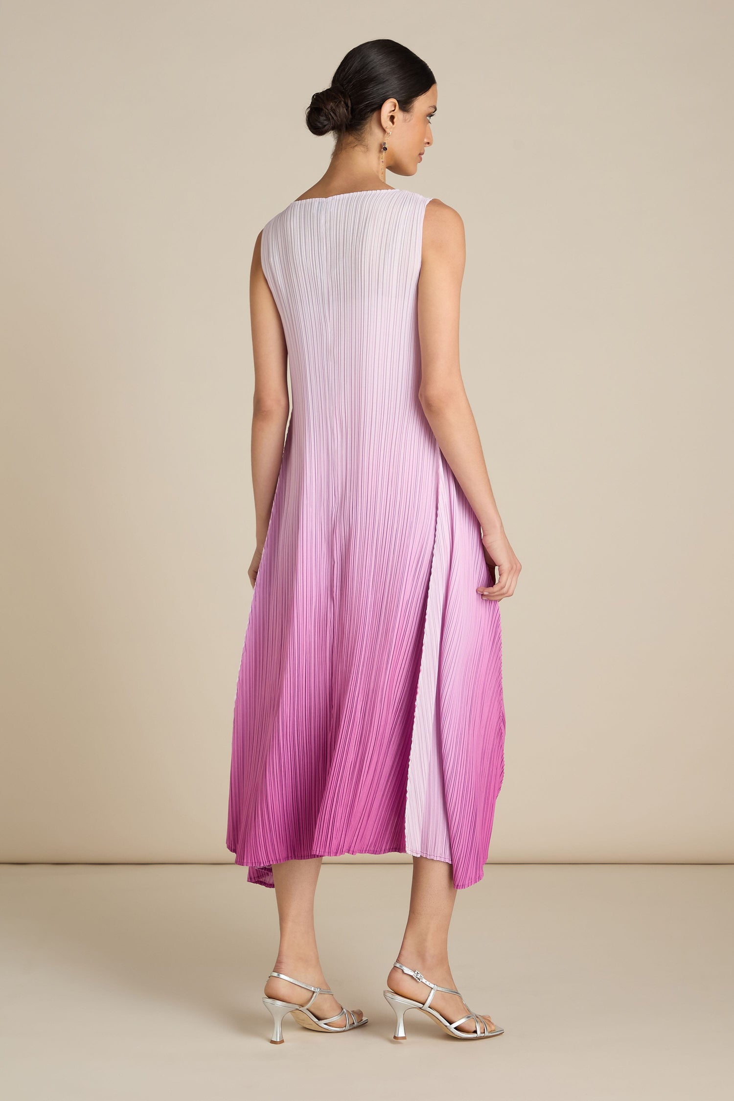 A woman stands with her back to the camera, wearing a sleeveless Pleated Dress that transitions from light purple at the top to deep pink at the bottom. The relaxed silhouette complements her bohemian flair, and she pairs it perfectly with silver high-heeled sandals.