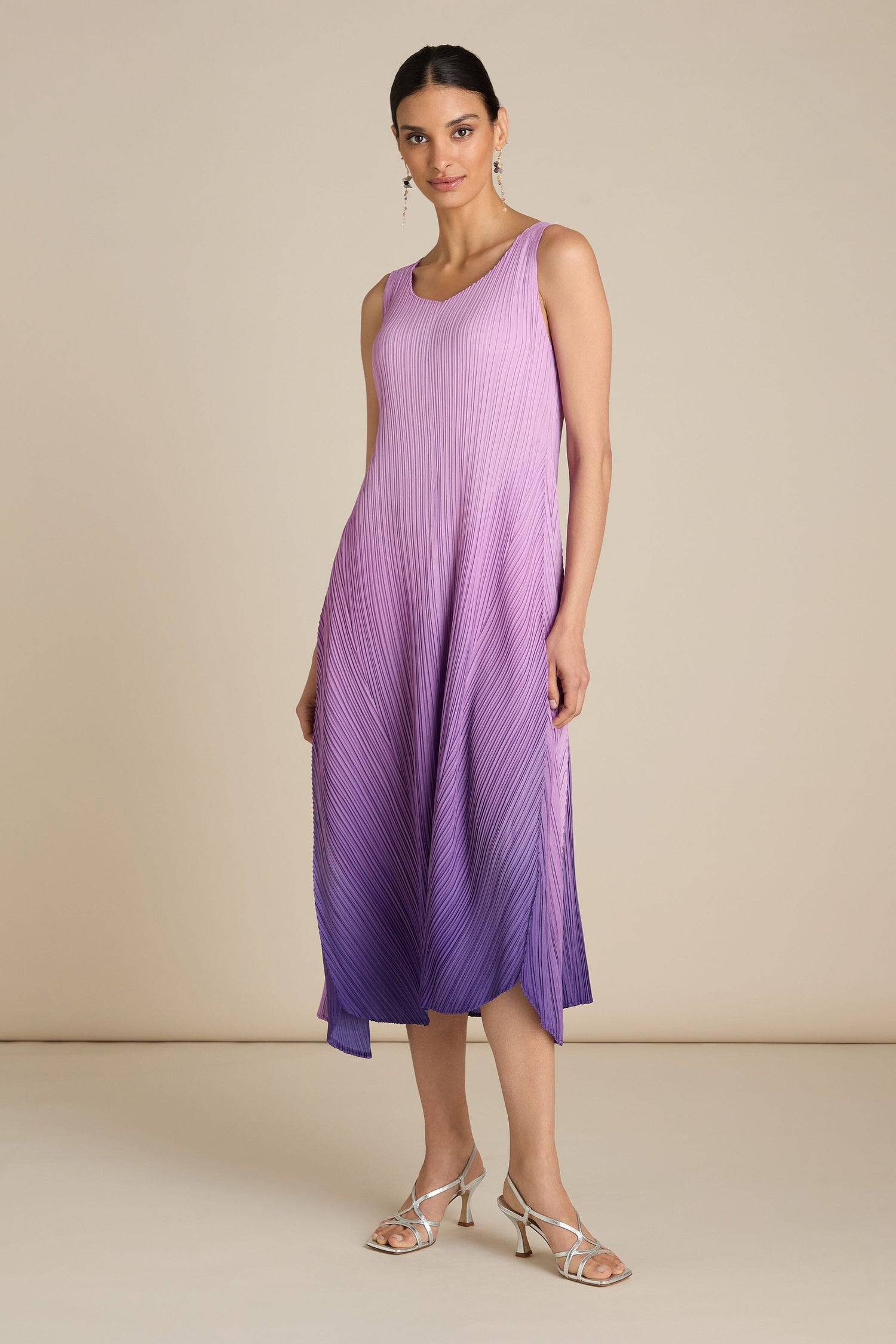 A woman stands against a plain background, wearing a sleeveless Pleated Dress in shades of purple. Paired with earrings and silver strappy heels, her outfit exudes a relaxed silhouette that hints at bohemian flair.