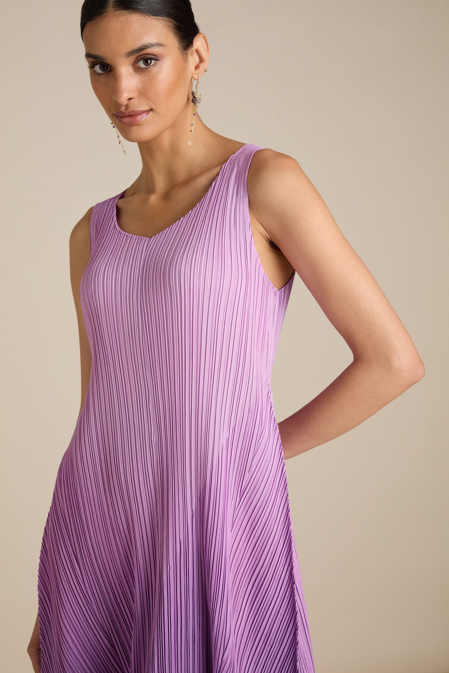 A woman wearing a Pleated Dress in lavender, featuring a sleeveless design and relaxed silhouette, stands against a neutral background with her right arm resting on her hip.