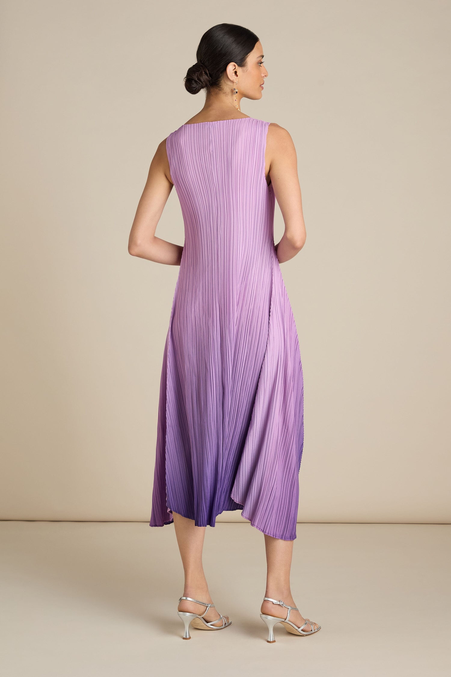 A woman wearing the Pleated Dress, which features gradient shades of purple and a sleeveless design, stands facing away with her hair styled in a bun and complemented by silver high heels. The relaxed silhouette of the dress adds a touch of bohemian flair to her elegant look.