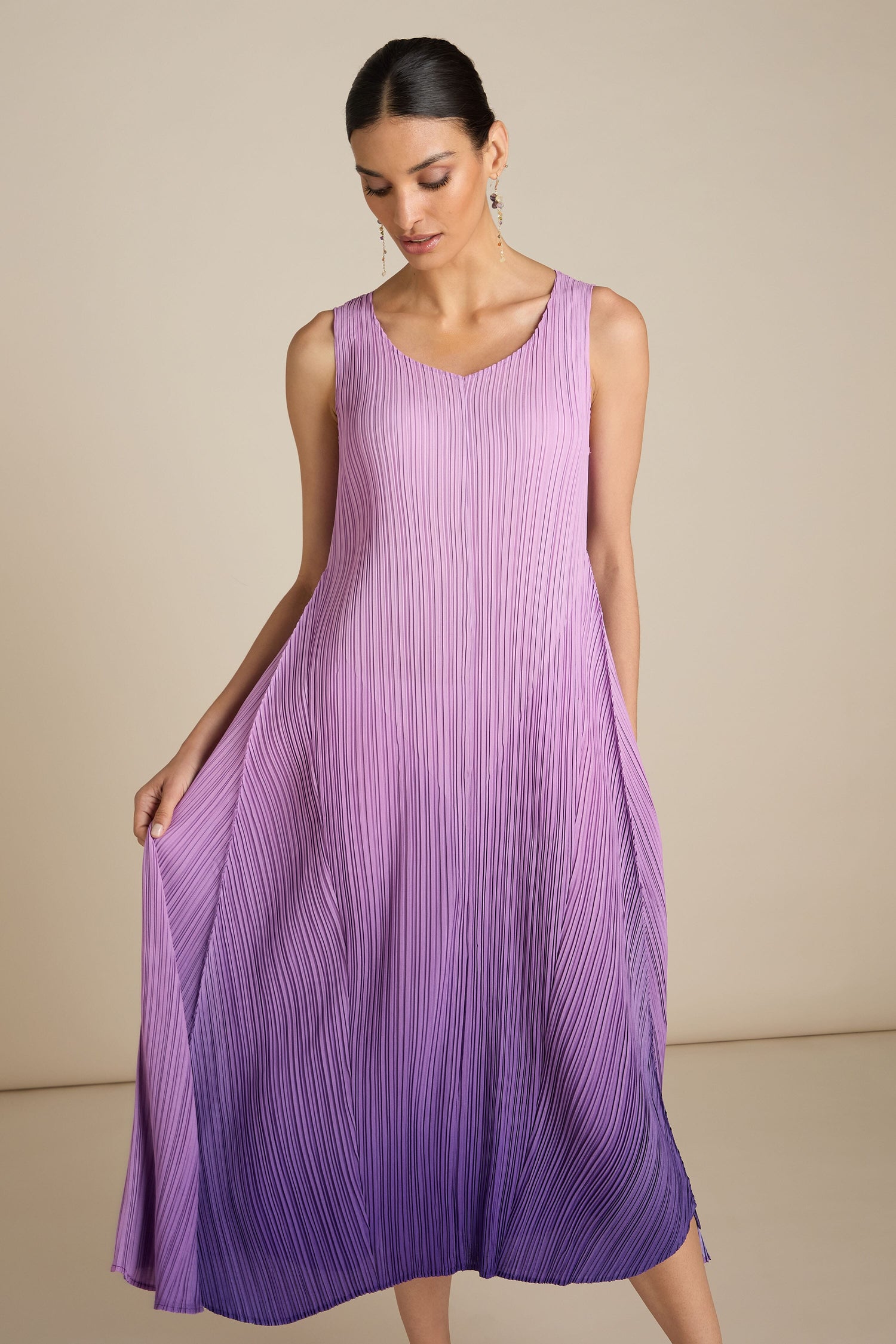 A woman in a purple gradient, sleeveless Pleated Dress looking down as she holds the sides of the dress, exuding a hint of bohemian charm.