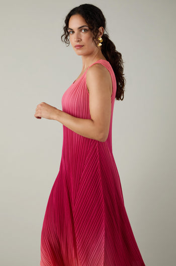 Woman with curly dark hair wearing gold earrings and a sleeveless Pleated Dress featuring shibori pleating in a pink-to-red ombre, standing against a plain light background.
