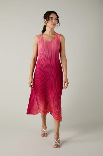 A woman in a pink, sleeveless Pleated Dress with shibori pleating and white strappy heels stands against a plain light background.