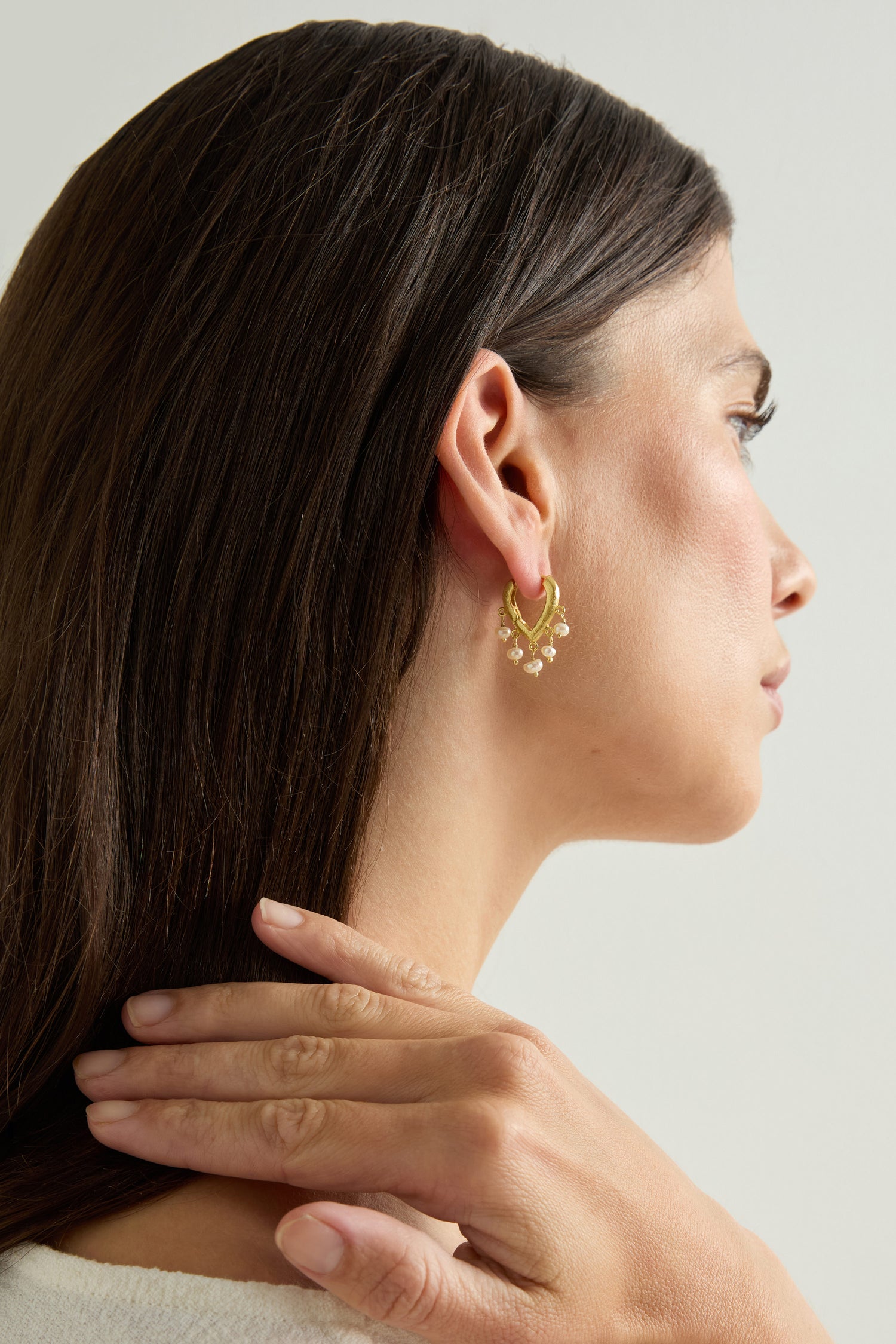A woman with long brown hair wears Gold Vermeil Pearl Teardrop Hoops, her hand resting on her shoulder, photographed from the side against a light background.