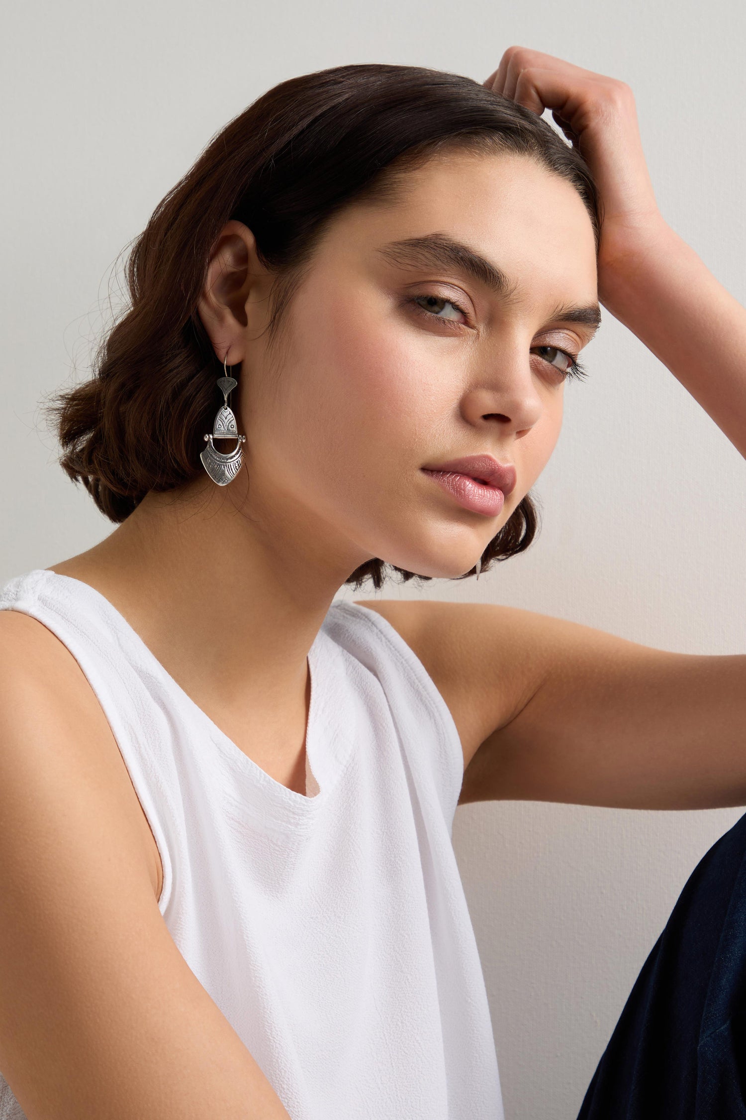 A person with short hair and a sleeveless white top sports Engraved Silver Tuareg Earrings, exuding elegance as they gaze neutrally at the camera, hand resting on their head.