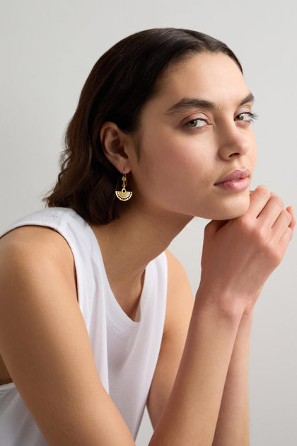 A person with short dark hair in a white sleeveless top rests their chin on their hands. Their Gold Vermeil Mini Fan Earrings add a touch of elegance to their look, being part of an exquisite jewellery collection.