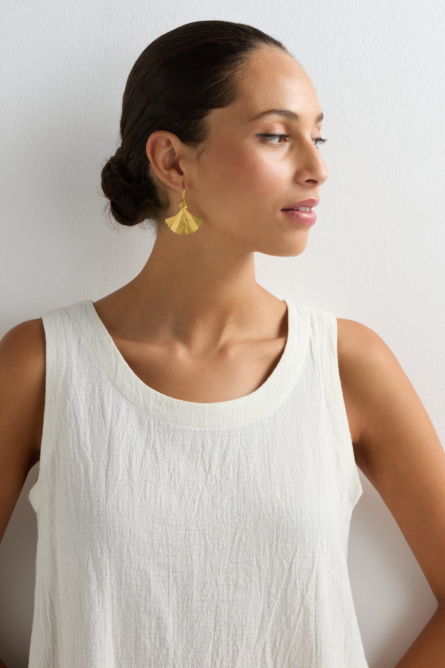 A woman with dark hair in a low bun wears a sleeveless white textured top and Gold Vermeil Ginkgo Leaf Earrings, standing against a white background and looking to the side, highlighting these elegant, nature-inspired earrings.