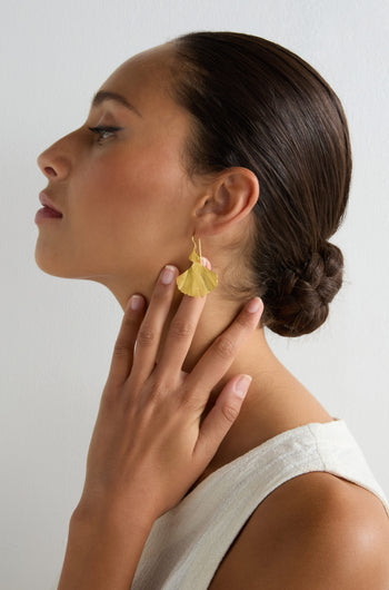 A woman with dark hair in a low bun wears a white top and Gold Vermeil Ginkgo Leaf Earrings, touching her neck with her hand against a plain light background.
