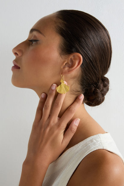 A woman with dark hair in a low bun wears a white top and Gold Vermeil Ginkgo Leaf Earrings, touching her neck with her hand against a plain light background.