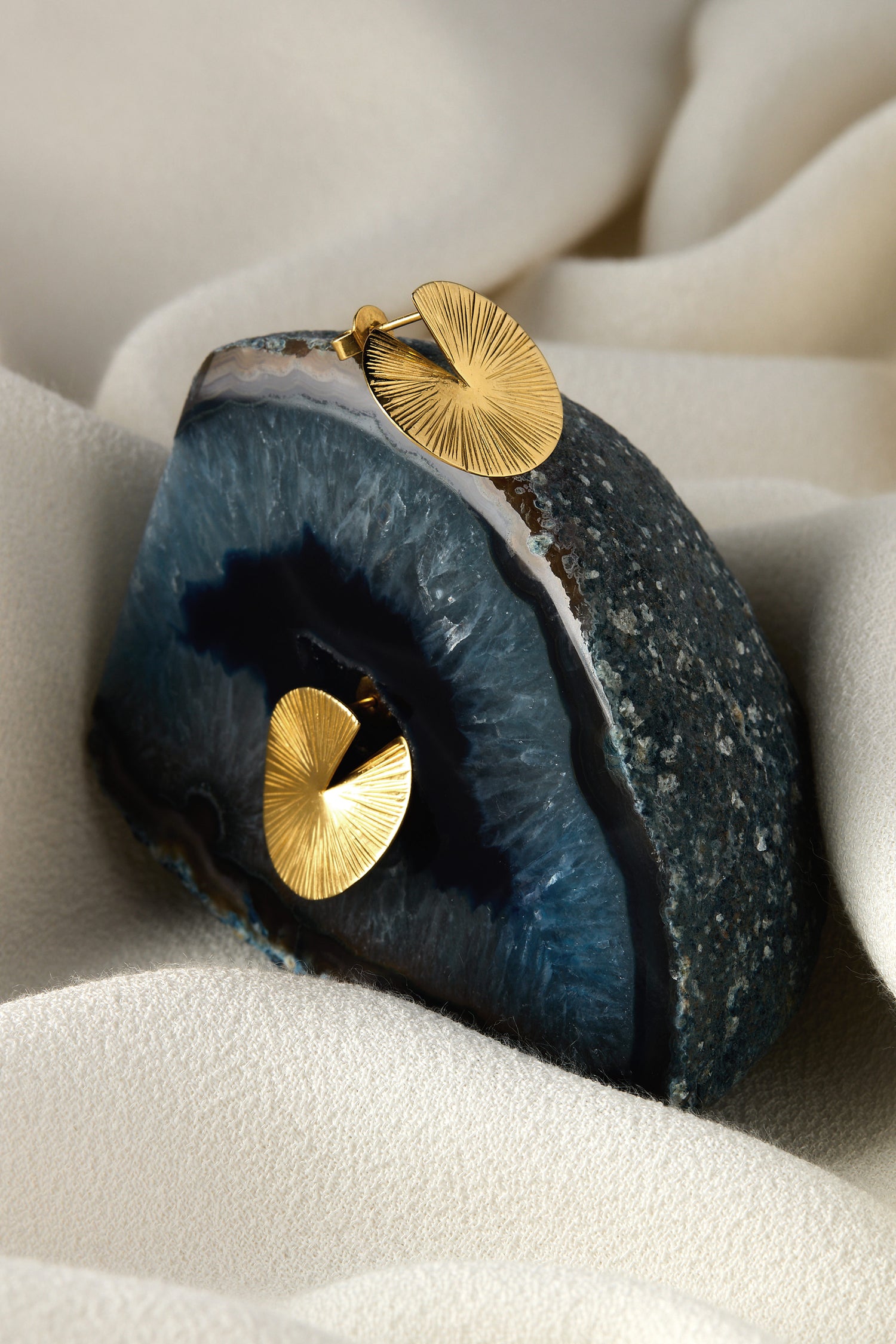 Small Gold Vermeil Lotus Leaf Earrings elegantly rest on a blue geode, highlighting their sculptural design against a textured beige fabric. The geode's natural veins contrast beautifully with the earrings' graceful silhouette.