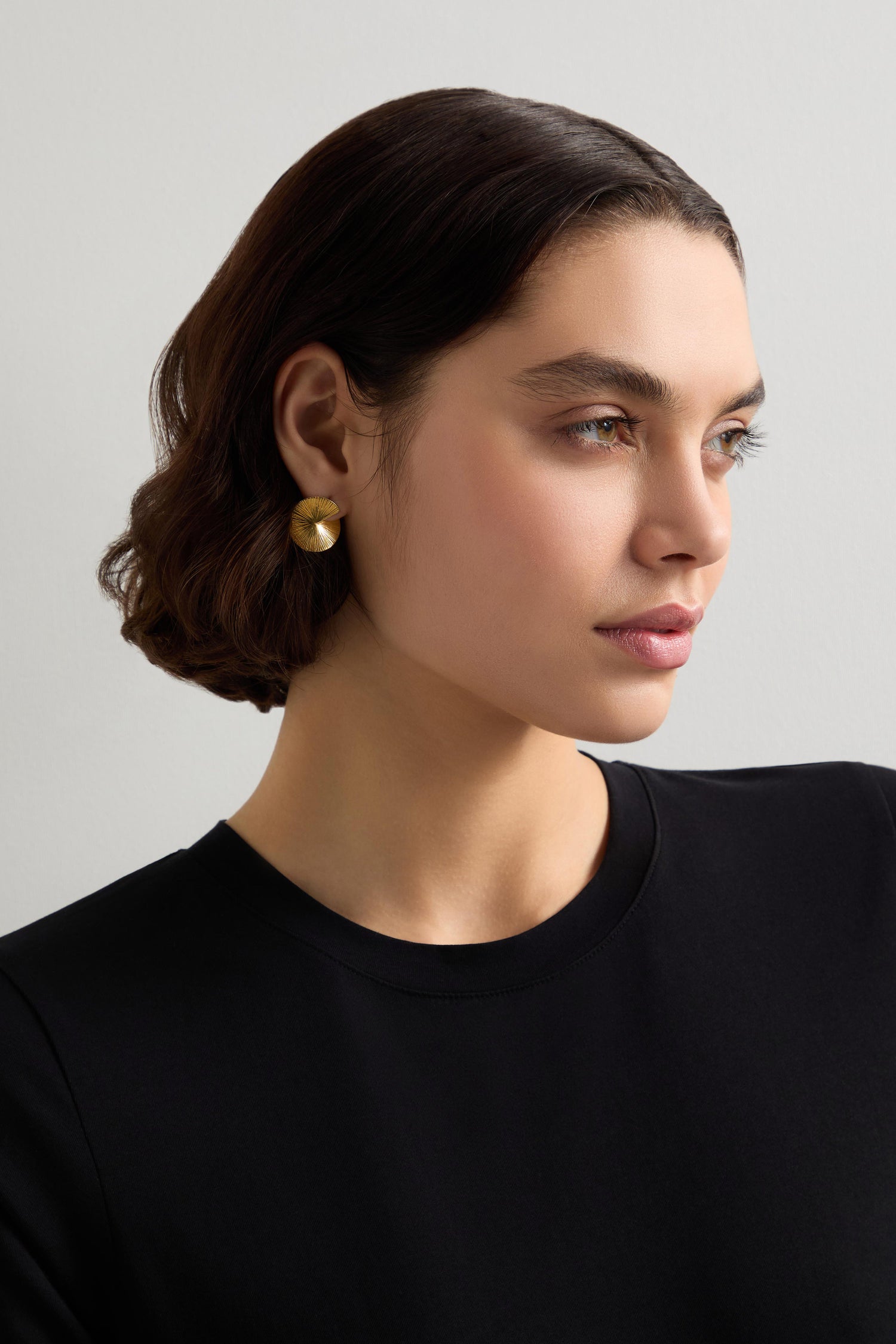 A person with short brown hair, wearing a black top and Small Gold Vermeil Lotus Leaf Earrings, gazes to the side against a neutral background.