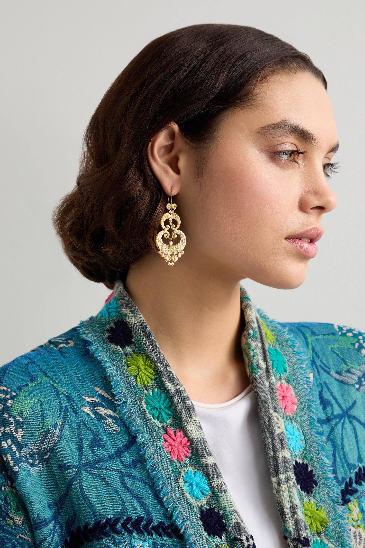 A woman with short brown hair wears Gold Vermeil Sumba Earrings adorned with traditional Sumbanese motifs. She pairs them elegantly with a blue floral-patterned jacket, adding an artisanal touch to her look.