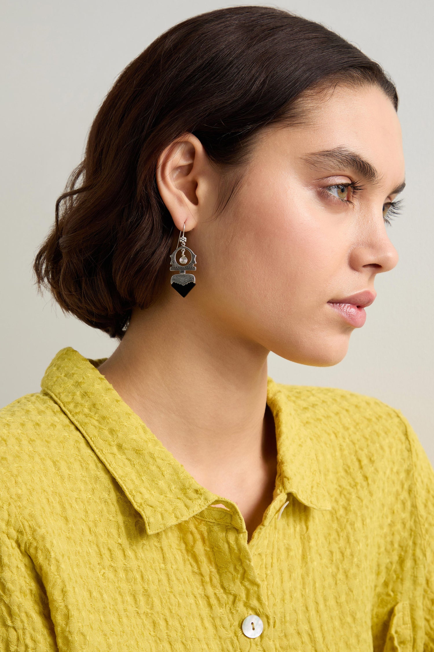 A person with short brown hair is wearing a yellow textured shirt and heart-shaped Silver and Black Onyx Tuareg Earrings, gazing to the side against a plain background.