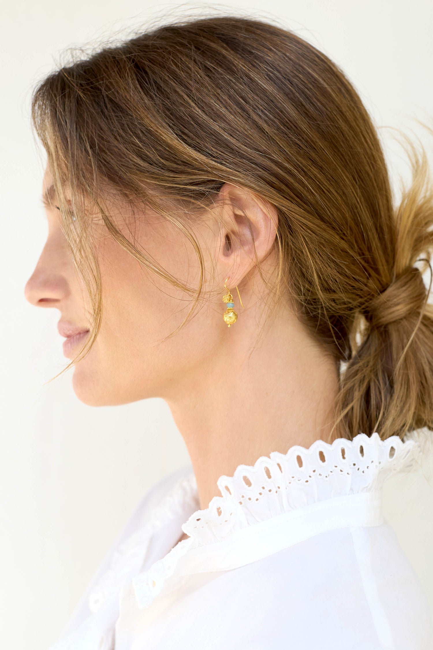 A woman with light brown hair in a low bun elegantly wears a white blouse with lace detailing, accentuated by striking Gold Vermeil Amazonite Bead Earrings from her jewelry collection.