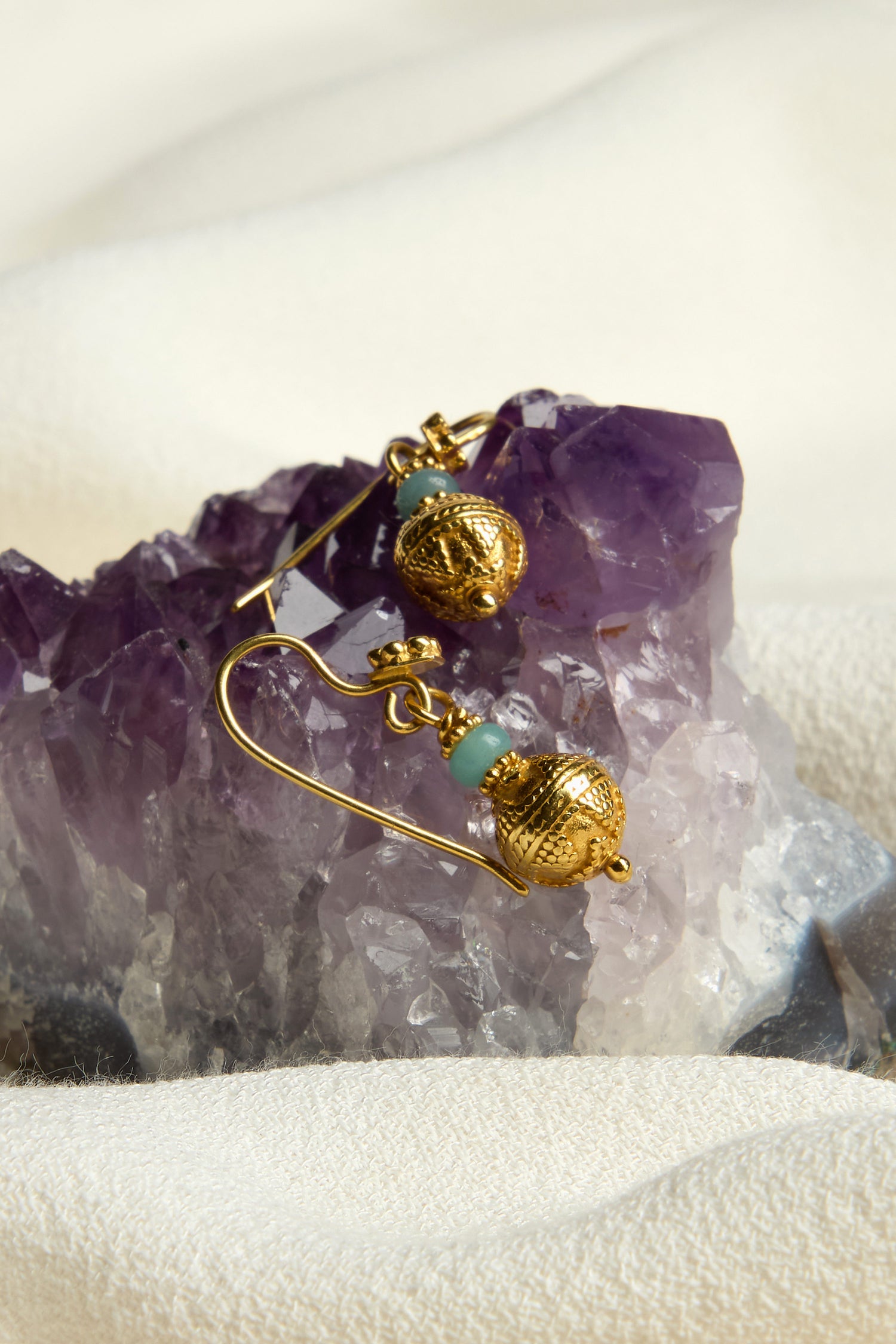 The Gold Vermeil Amazonite Bead Earrings, showcasing spherical designs and vibrant turquoise beads, elegantly rest on an amethyst crystal against a soft white fabric background, epitomizing the sophistication of our exclusive jewelry collection.