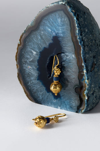 Against a neutral background, a pair of Gold Vermeil Lapis Bead Earrings is displayed: one hangs on a blue geode, and the other lies in front.