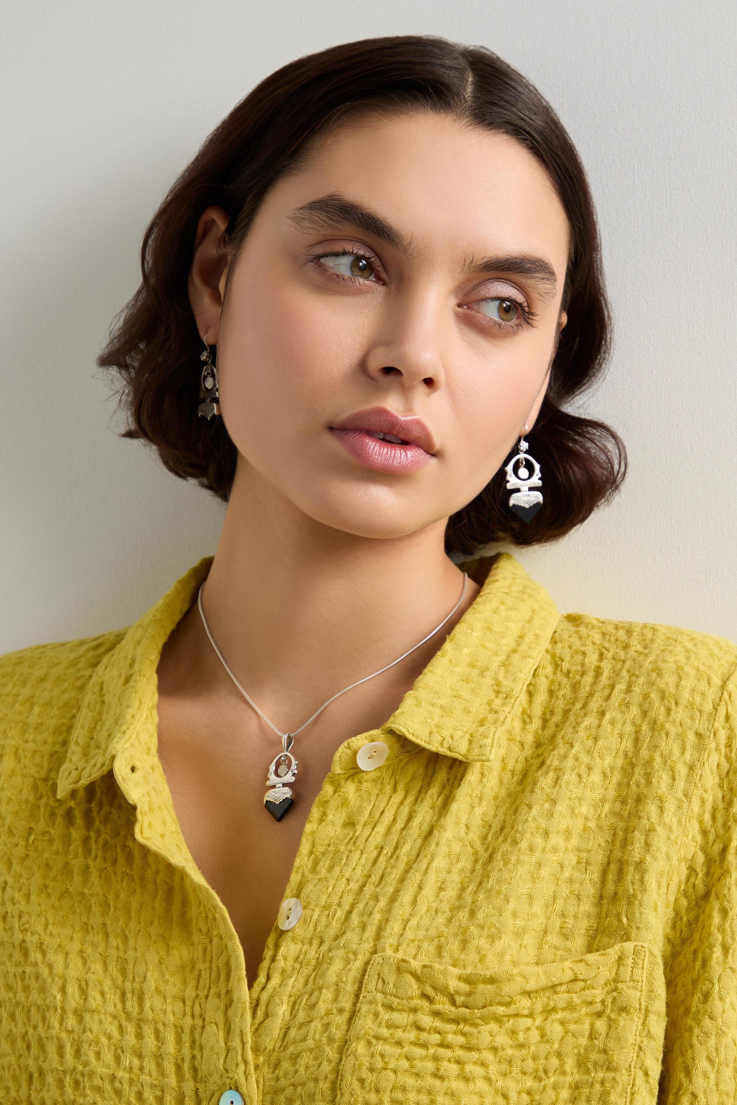 A person with short dark hair wears a yellow textured shirt, complemented by a Tuareg Silver and Black Onyx Pendant along with black and silver jewelry, gazing to the side against a plain background.