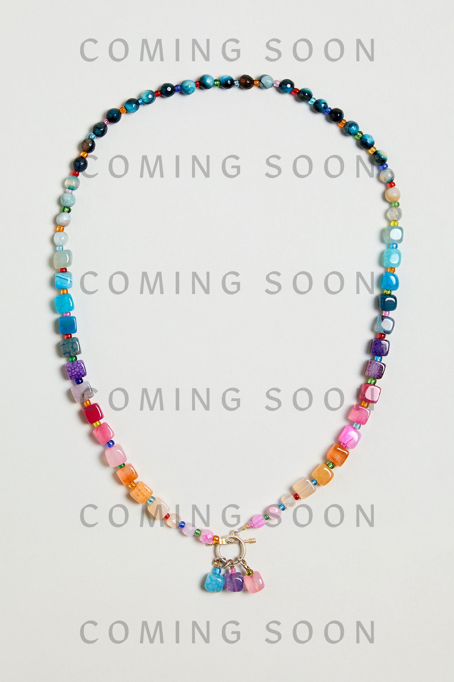 The Artisanal Rainbow Agate Toggle Necklace features vibrant beads, a gold clasp, and dangling charms. Part of the ARBAGM6NLA collection, it’s shown on a plain background with "COMING SOON" text overlaid.