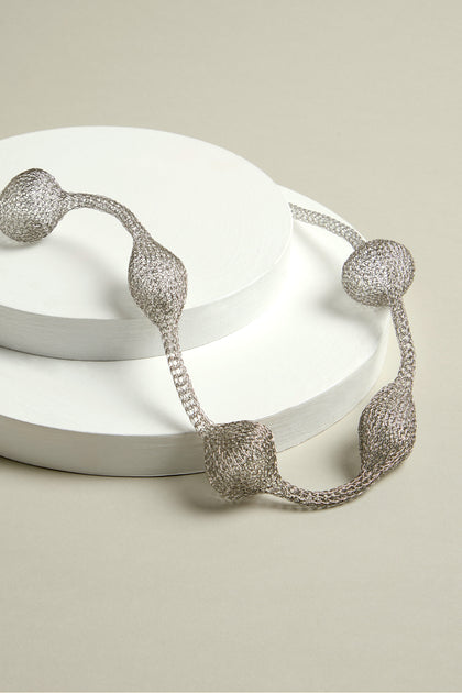 The Hand Crocheted Orb Necklace, with its rounded segments, exudes an elegant balance as it rests gracefully on two white circular platforms, resembling metallic orbs.