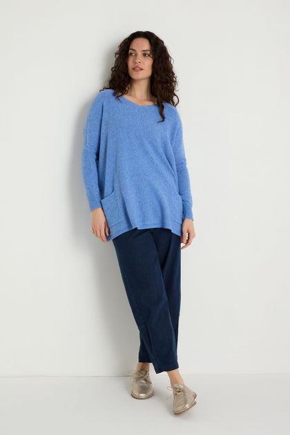 A person stands against a white wall wearing the Two Pocket Oversize Knit in blue, paired with dark blue pants and beige shoes—a perfect example of cozy women's knitwear.